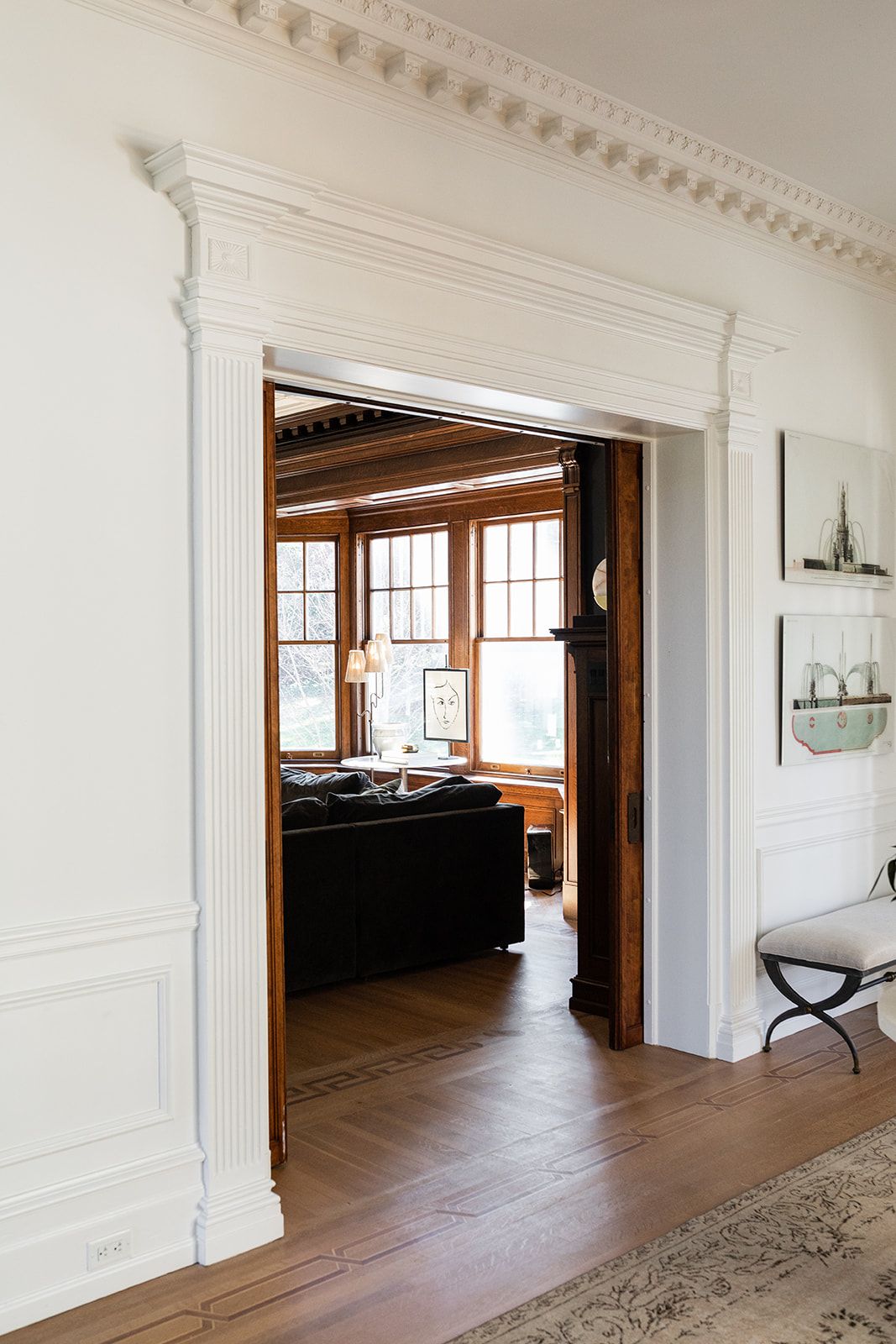 White doorway with crown molding leads to sunlit room with hardwood floors and dark furniture.