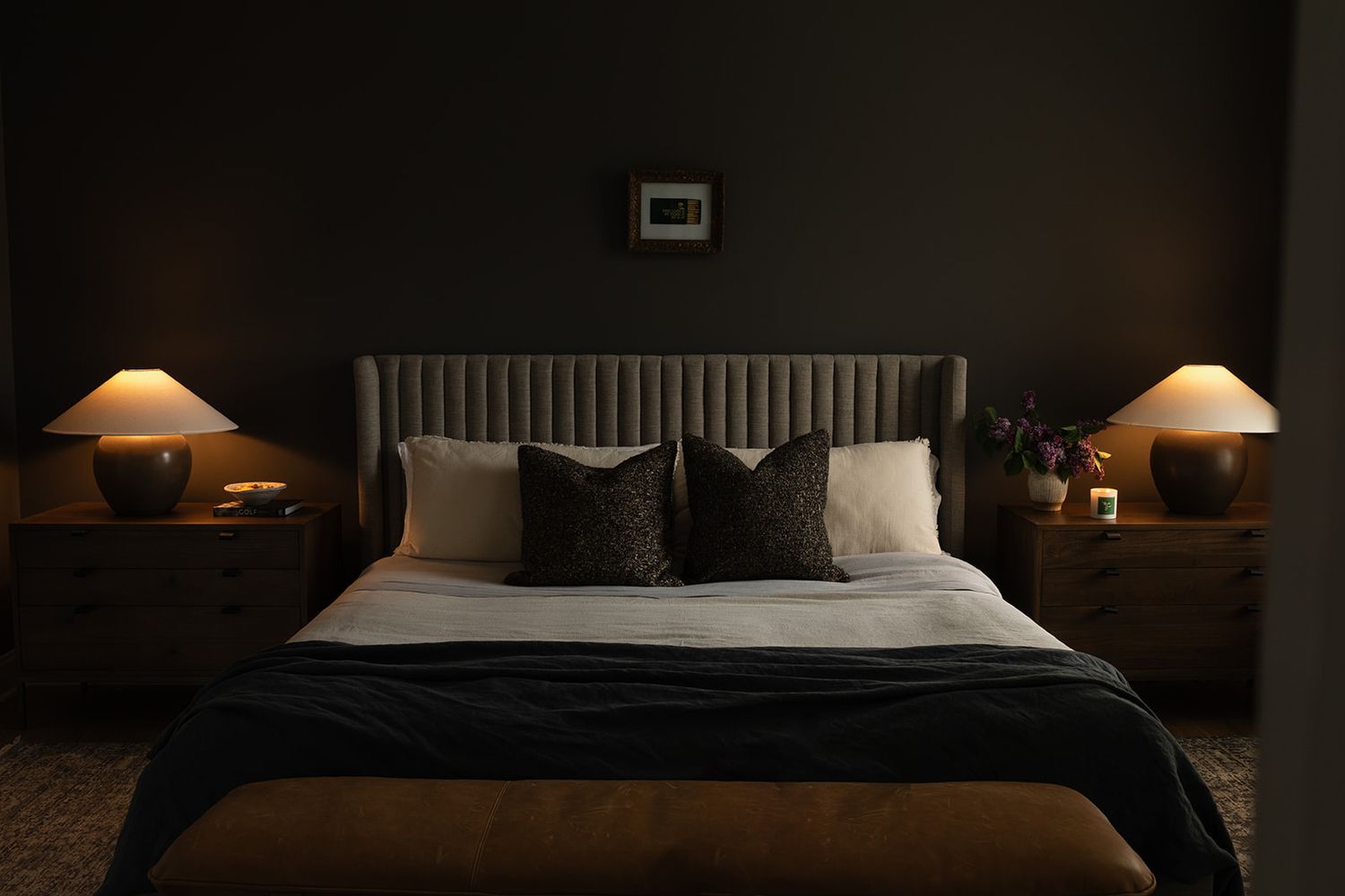 A cozy dark bedroom with a bed featuring white linens, black pillows, and matching bedside lamps with warm lighting.