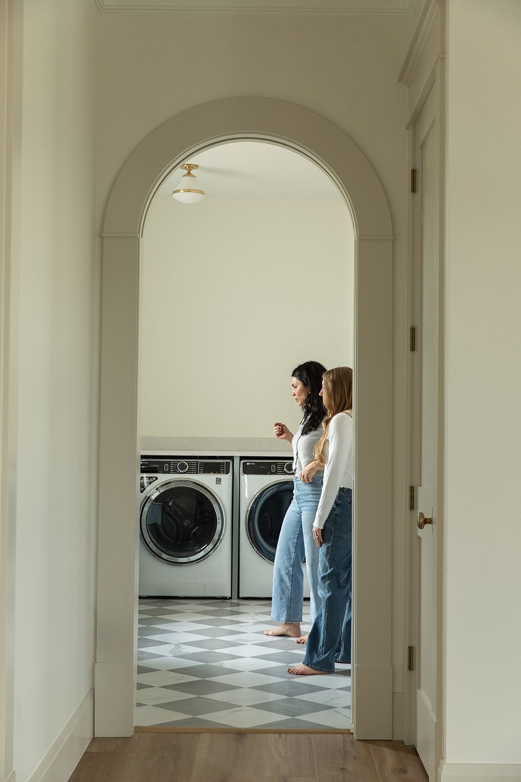 Two women stand in a bright laundry room with checkered floors and modern washing machines framed by an elegant arch.