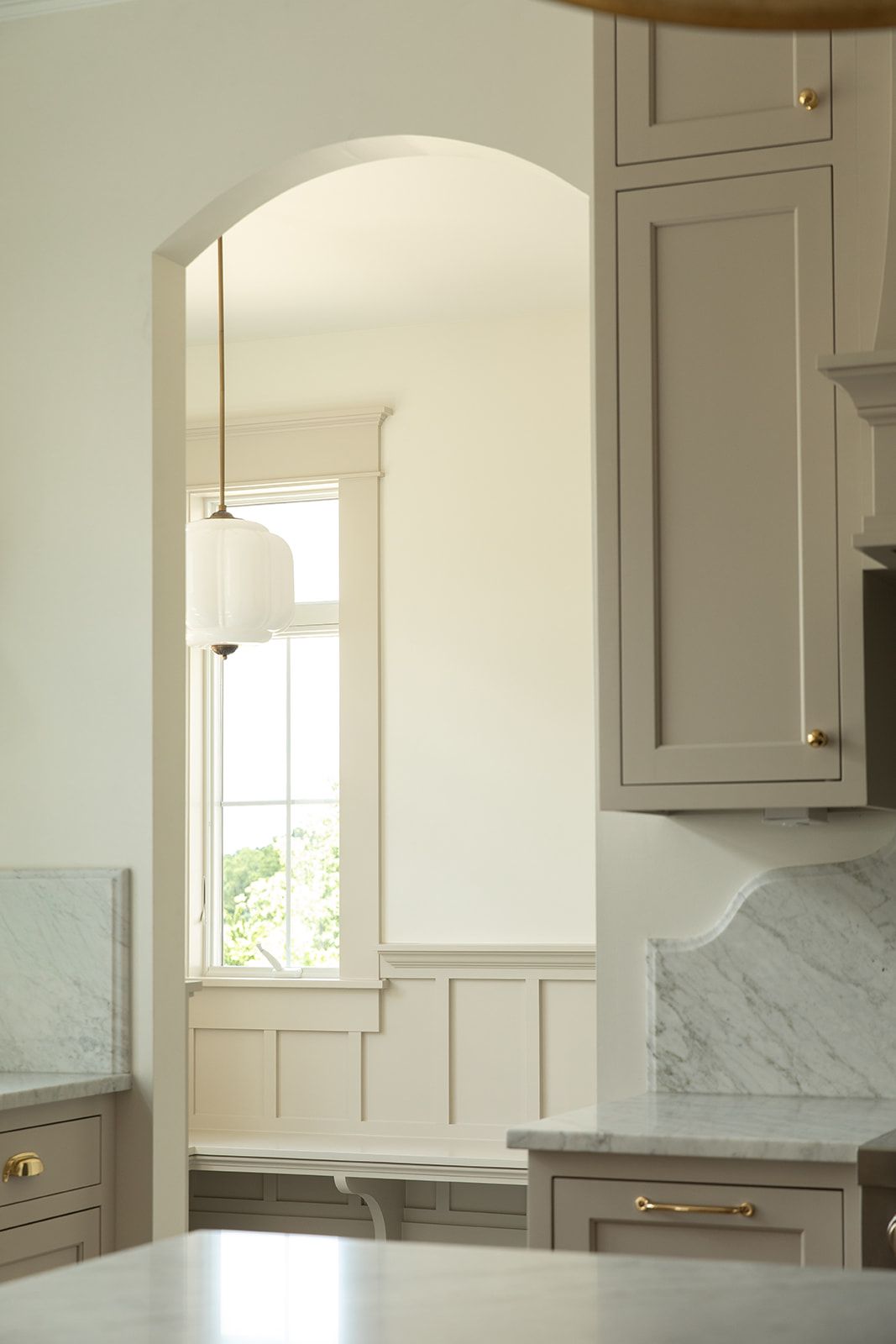 Elegant kitchen with greige cabinets, marble countertops, brass hardware, arched doorway, and a white pendant light over a window.