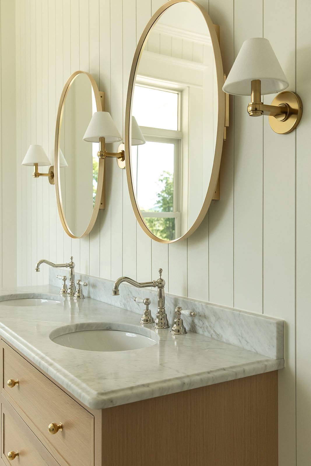 Elegant double vanity with marble countertop, oval gold mirrors, brass sconces, and wood cabinets in a bright bathroom.