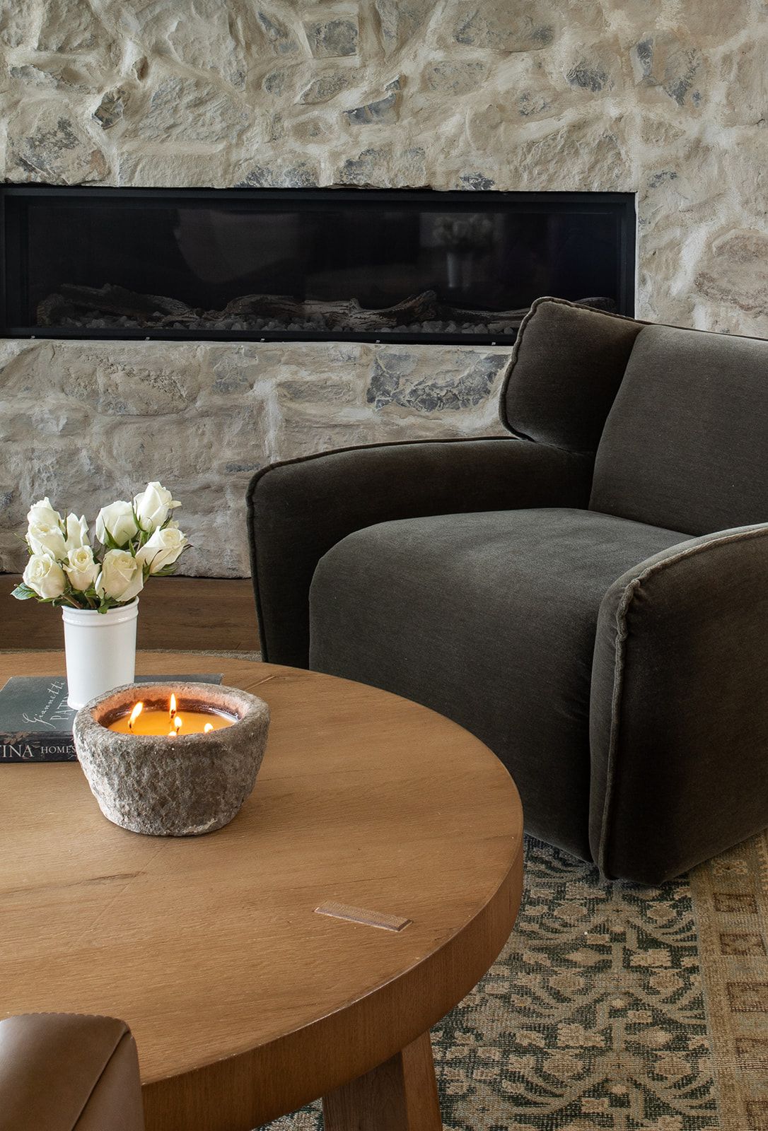 Cozy living room with olive velvet armchair, round wood coffee table, lit candle, white roses, and stone fireplace backdrop.