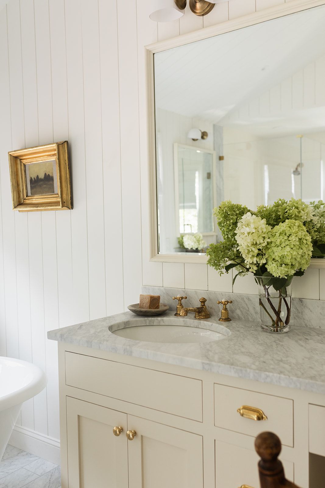 Elegant bathroom vanity with gray marble countertop, brass fixtures, hydrangea arrangement, and gold-framed artwork.