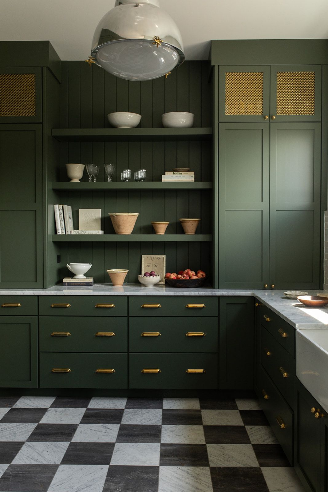 Dark green kitchen with gold hardware, open shelving, marble countertops, and black-and-white checkered floor tiles.
