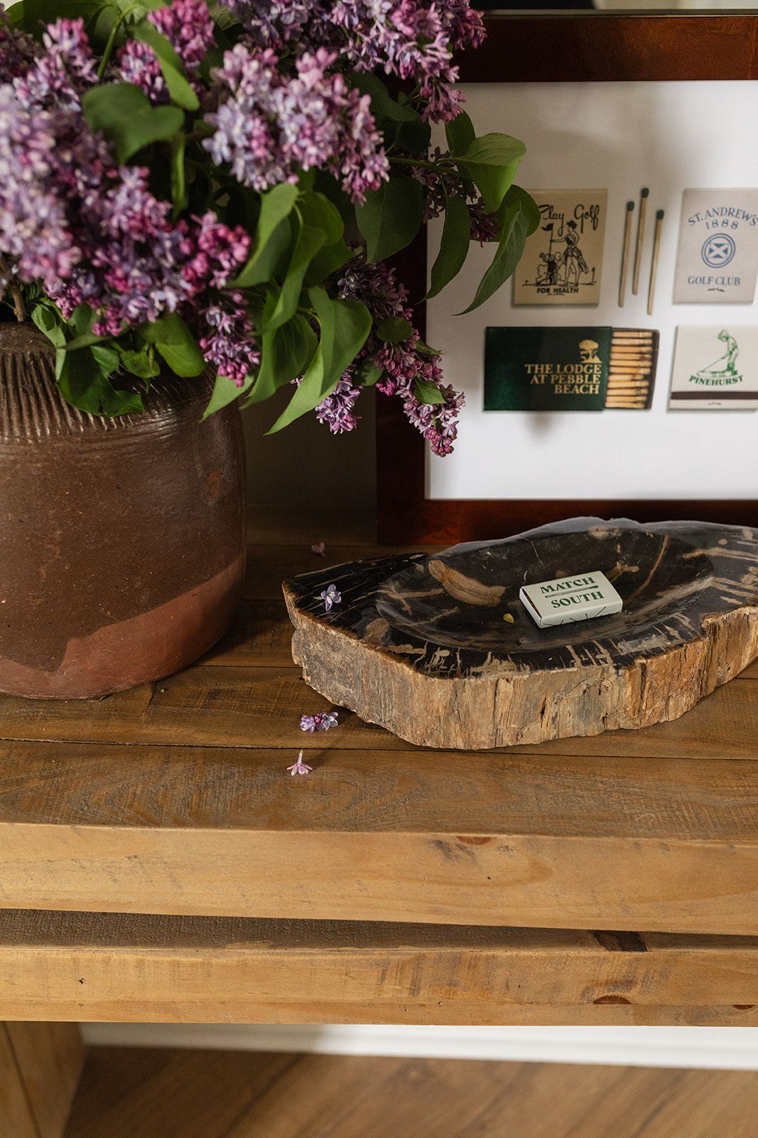 A rustic wooden table displays a potted lilac plant and a wooden slice coaster, with matches visible in the background.