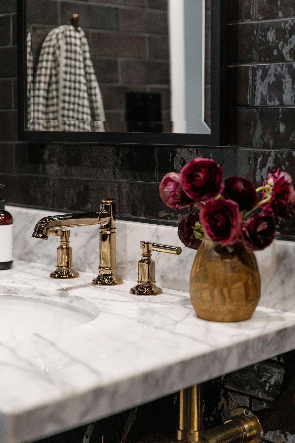 Elegant bathroom vanity with gold faucets, marble countertop, black walls and burgundy flowers in a ceramic vase.