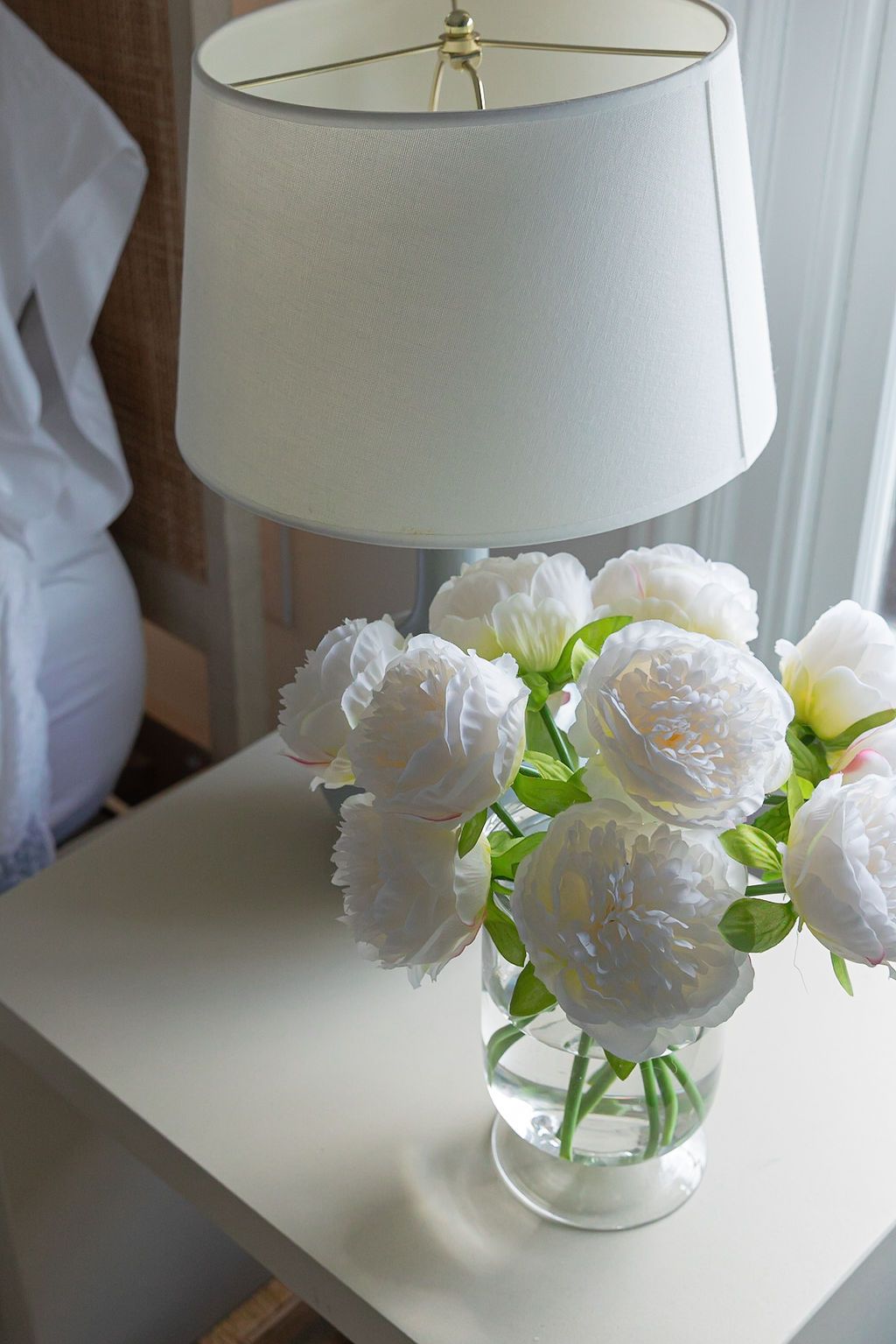 White peonies arranged in a glass vase under a classic drum lampshade create an elegant vignette.