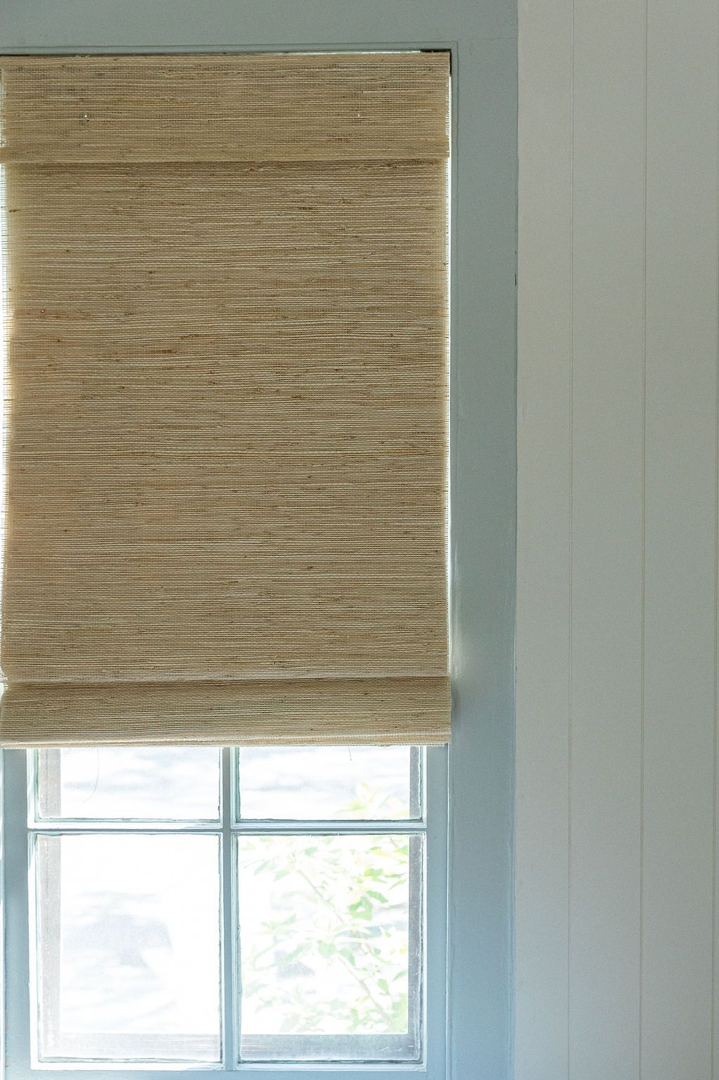 Natural woven bamboo window shade in beige hangs over a white-framed window with light filtering through.