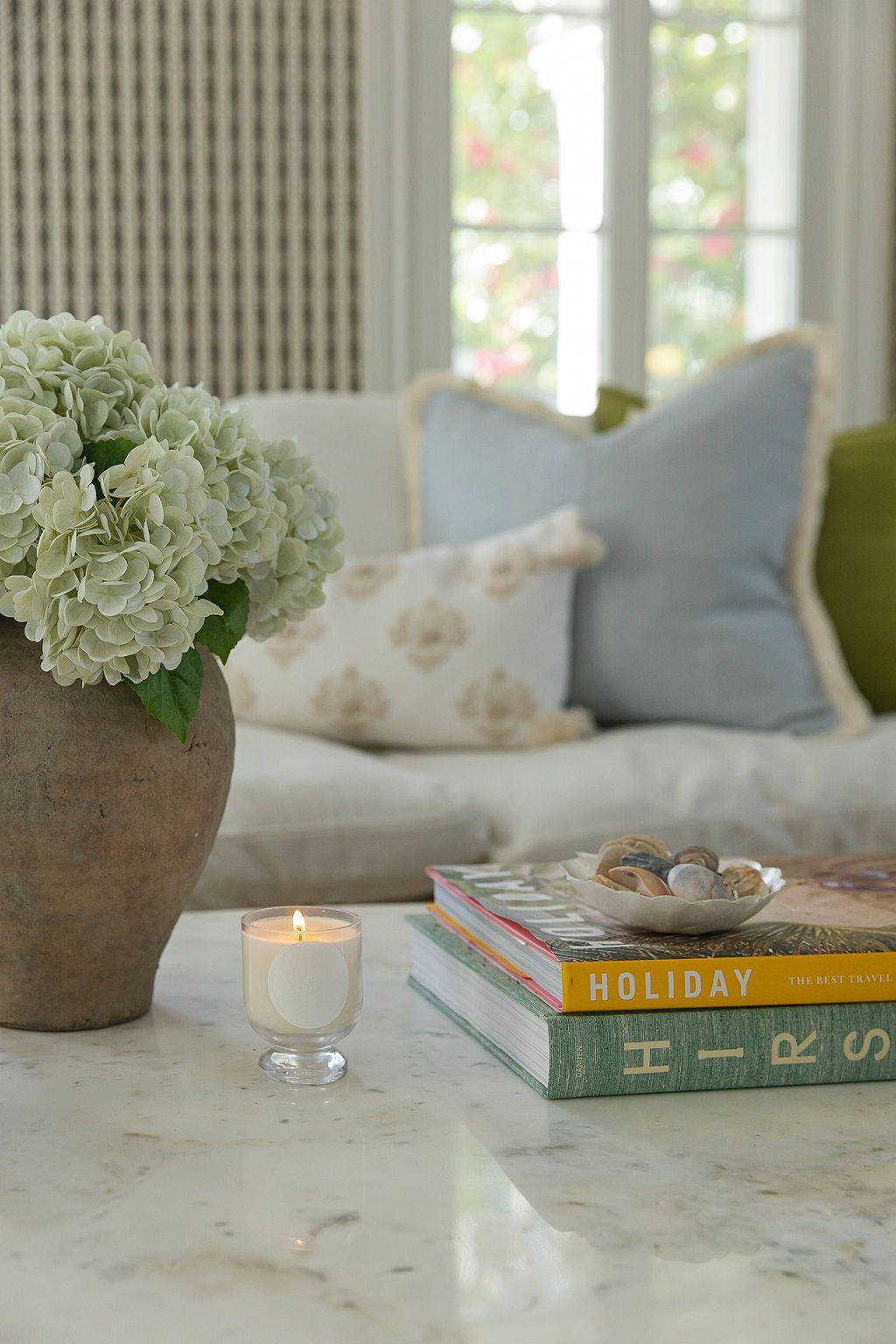 White hydrangea flowers in a stone pot next to stacked books and a lit candle on a marble surface.