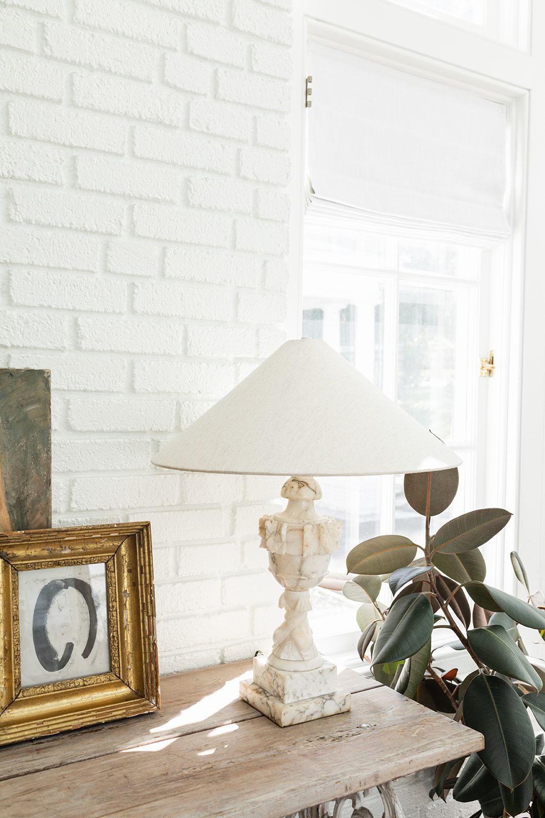 White table lamp and framed artwork arranged on vintage console table against white brick wall.