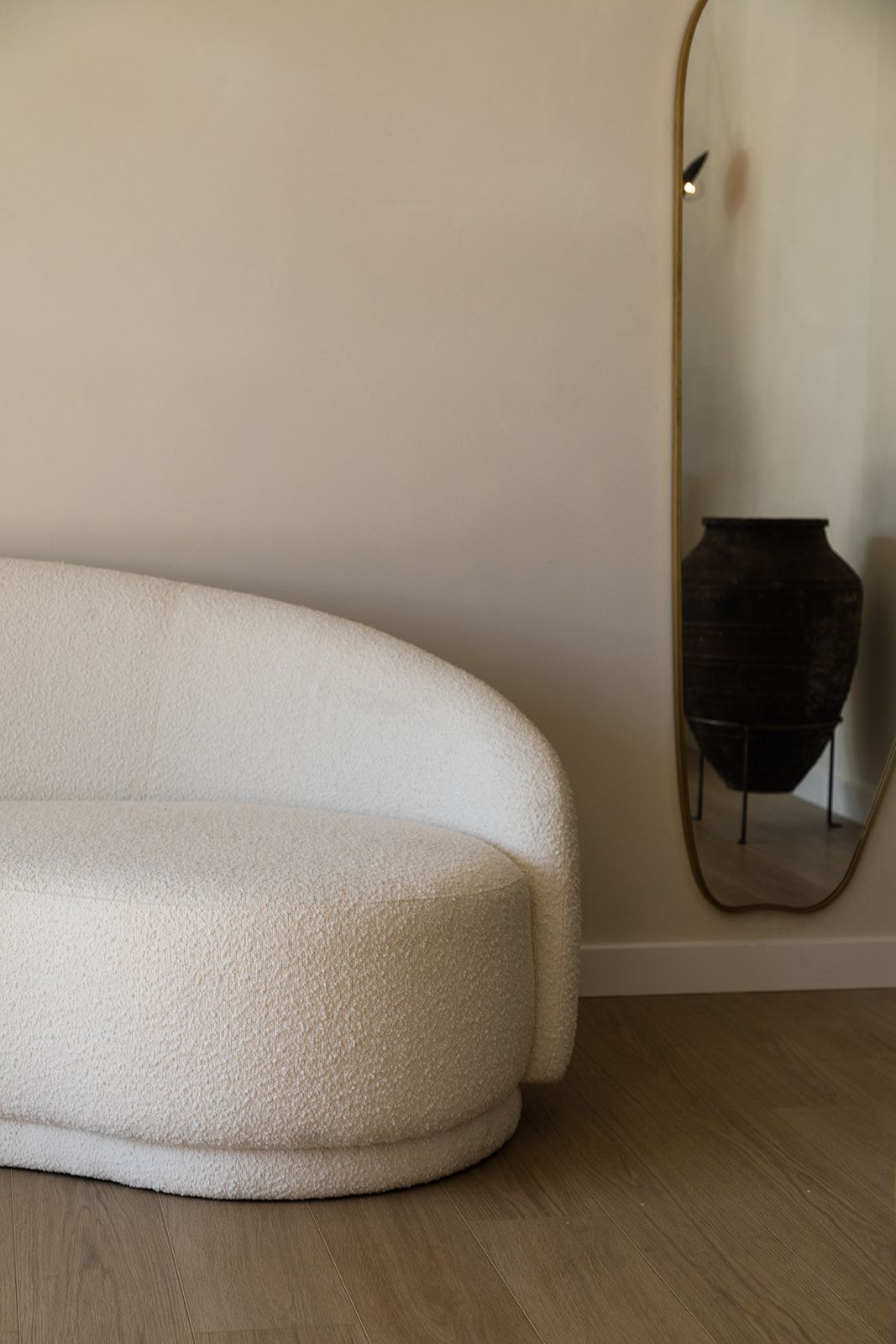 Curved white boucle sofa against beige wall with arched mirror reflecting minimalist interior design.