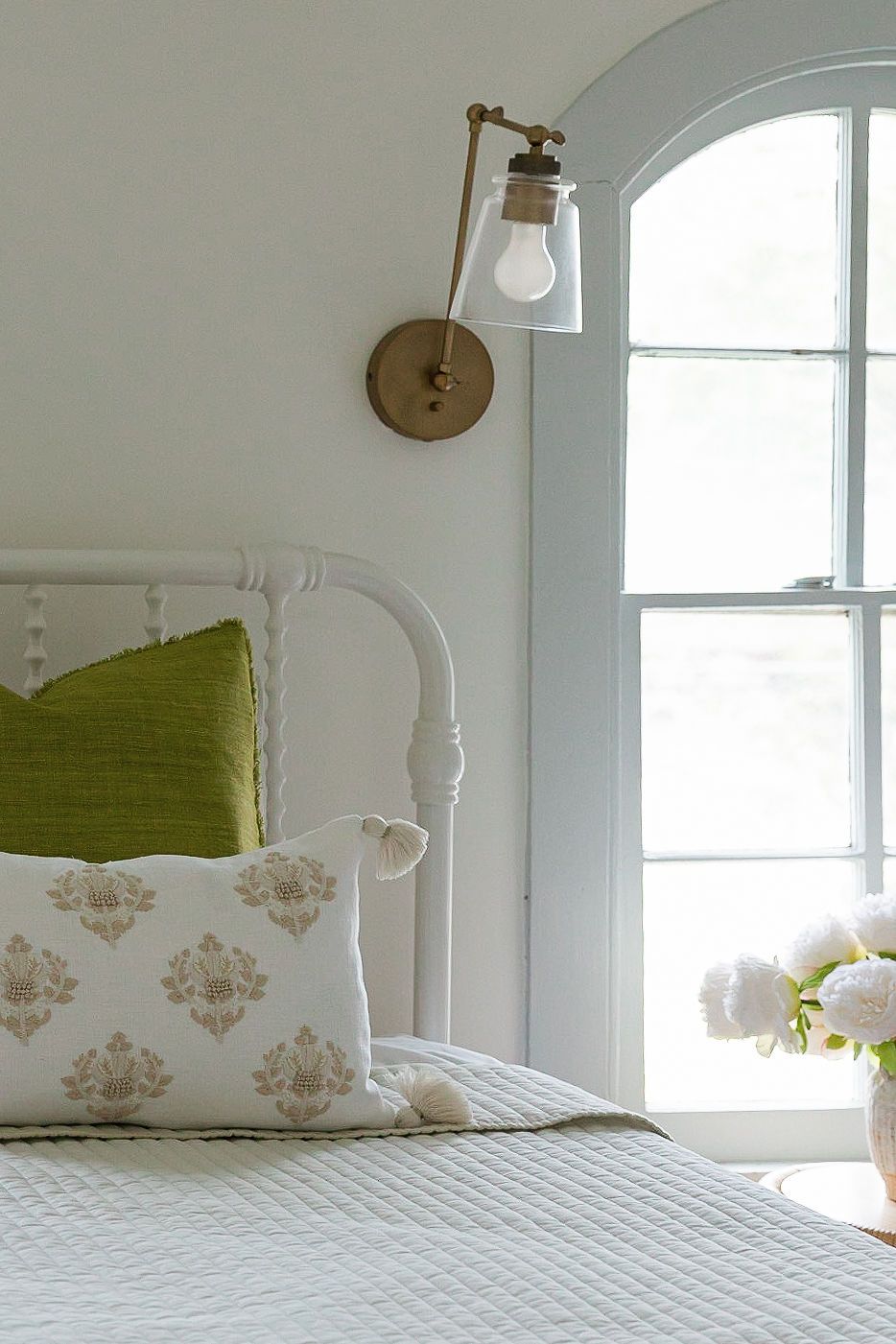 Bright bedroom interior featuring white bedding, green accent pillows and arched window with wall sconces.