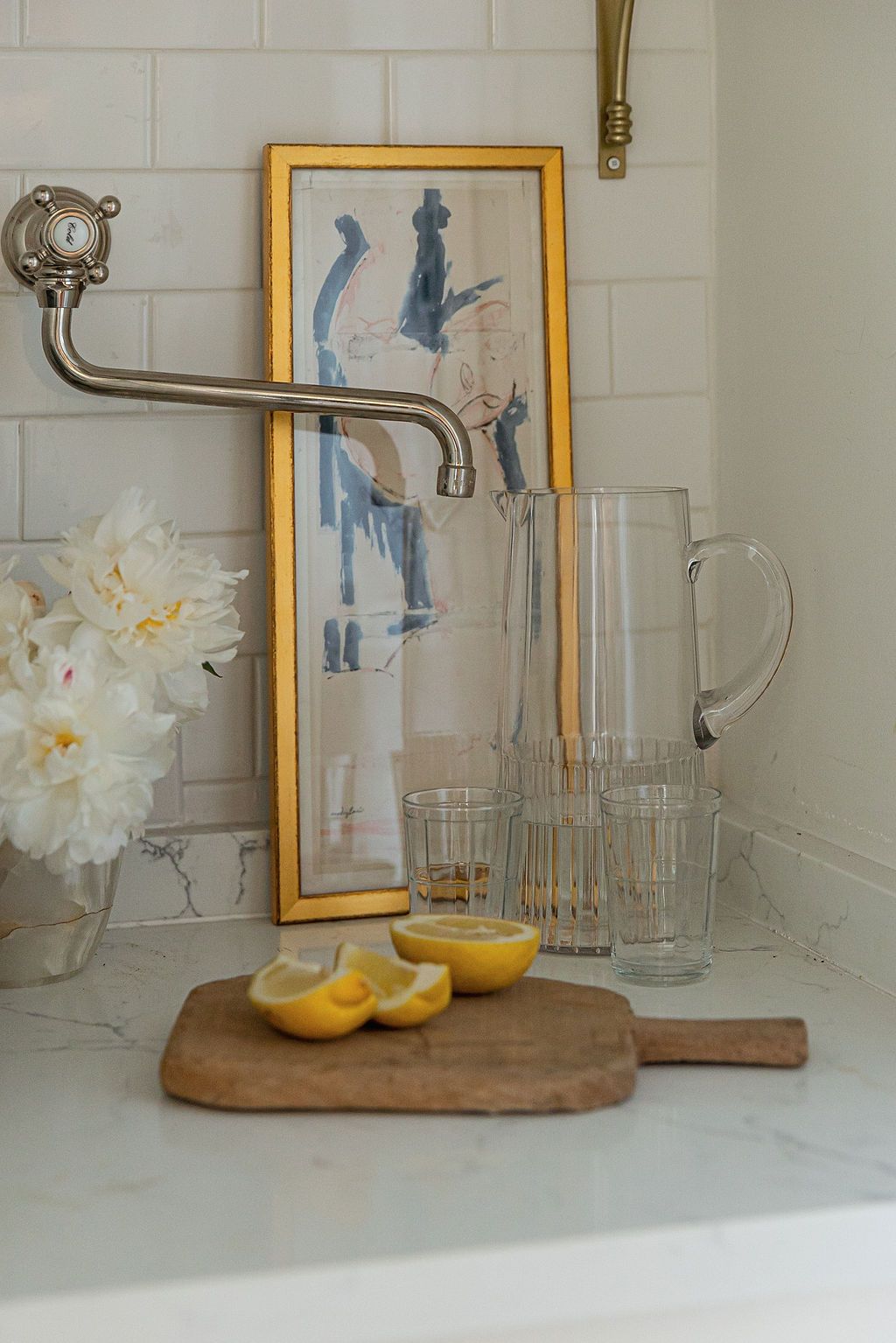 Wooden cutting board with lemons and floral decor on white countertop with decorative framed artwork in background.