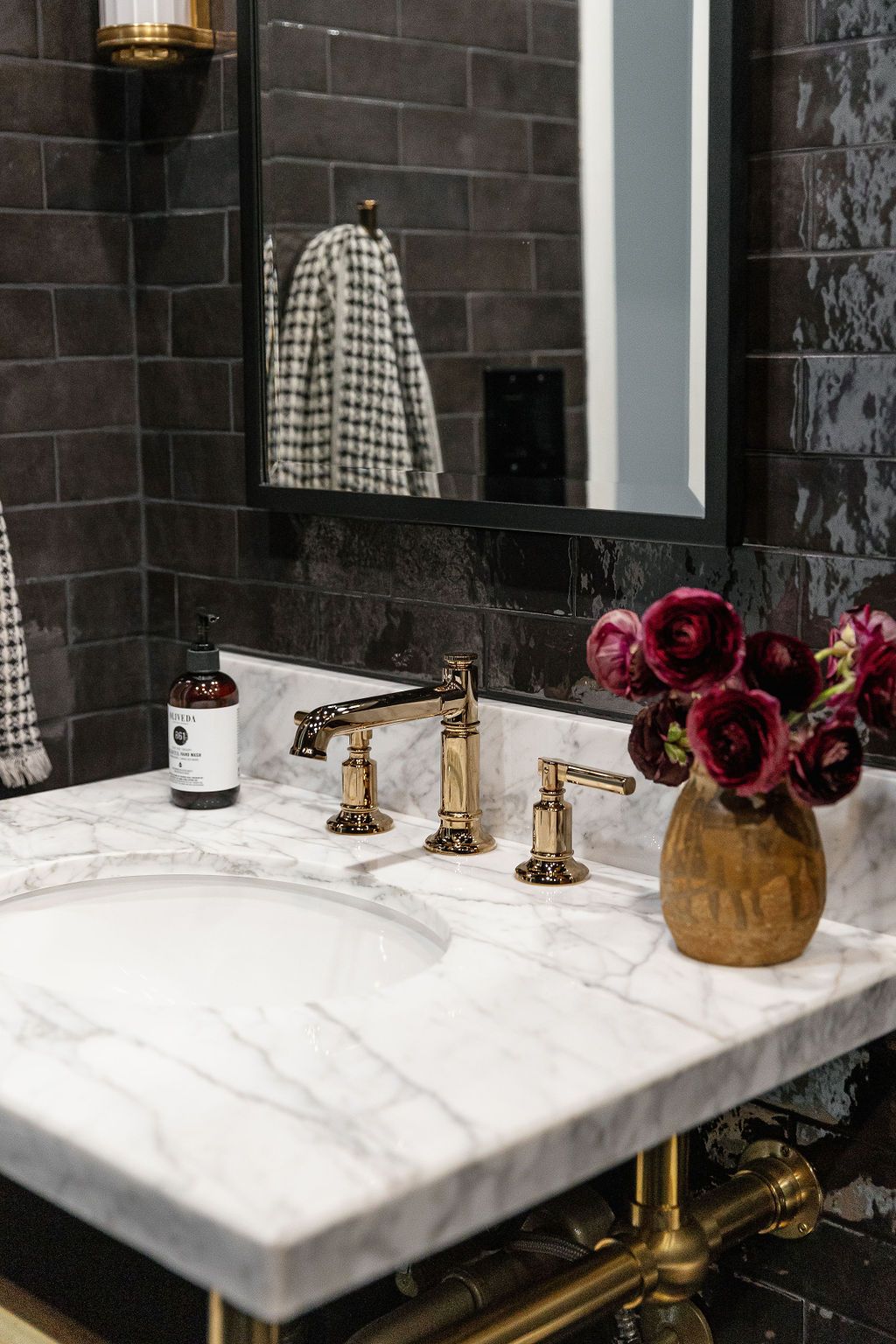 Luxurious bathroom vanity with marble countertop, brass fixtures, dark tile walls, and burgundy flowers in a gold vase.