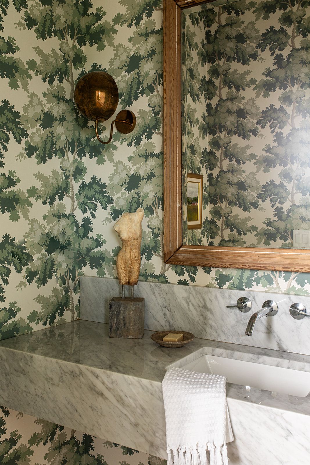 Elegant bathroom with green leaf wallpaper, marble sink, gold mirror, brass sconce, and decorative torso sculpture.