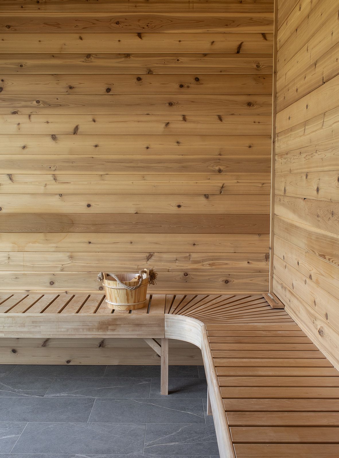 Minimalist cedar wood sauna interior with slatted benches, slate tile floor, and a wooden bucket in a corner.