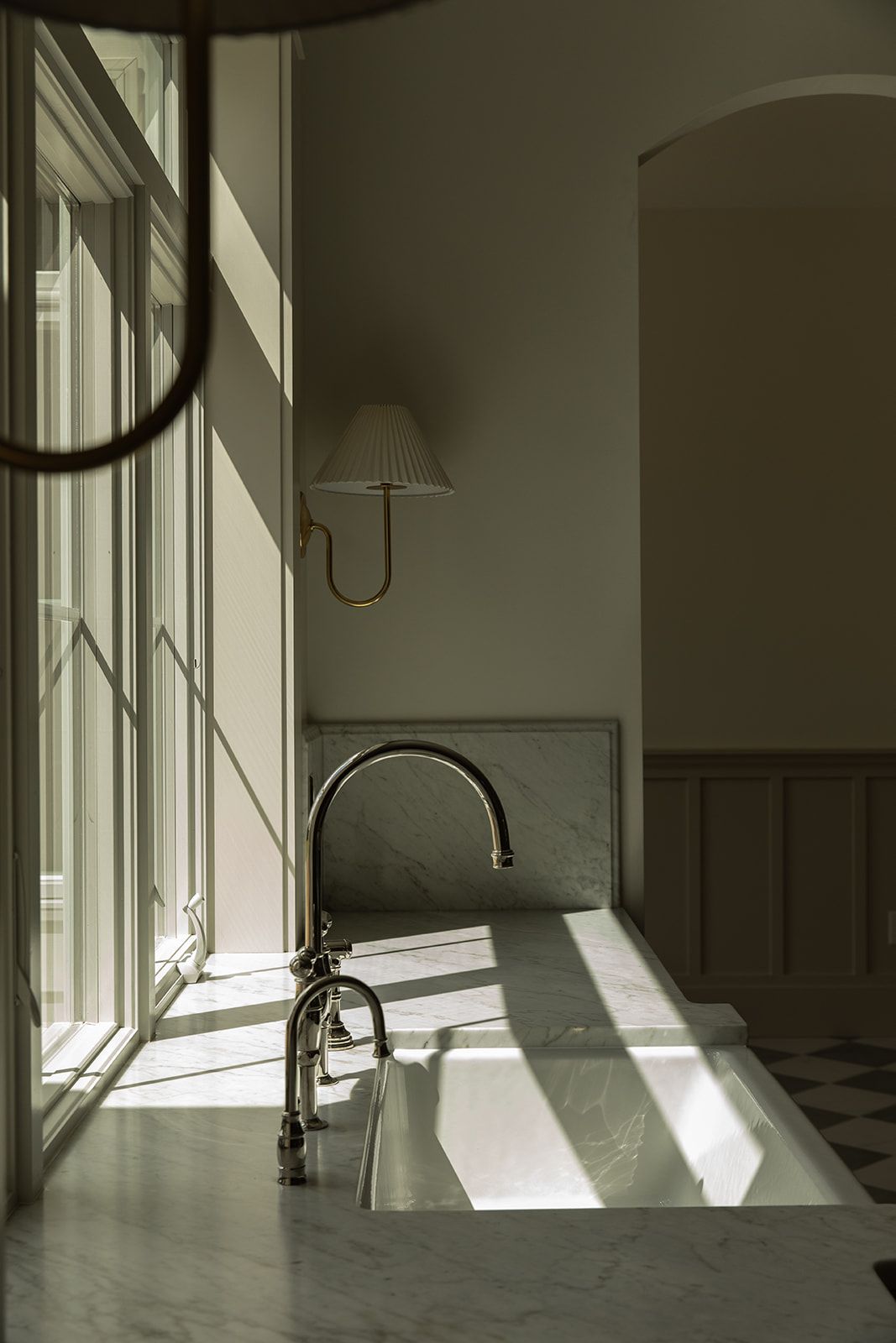 Sunlit marble kitchen sink with polished nickel gooseneck faucet, pleated wall sconce, and arched doorway in background.