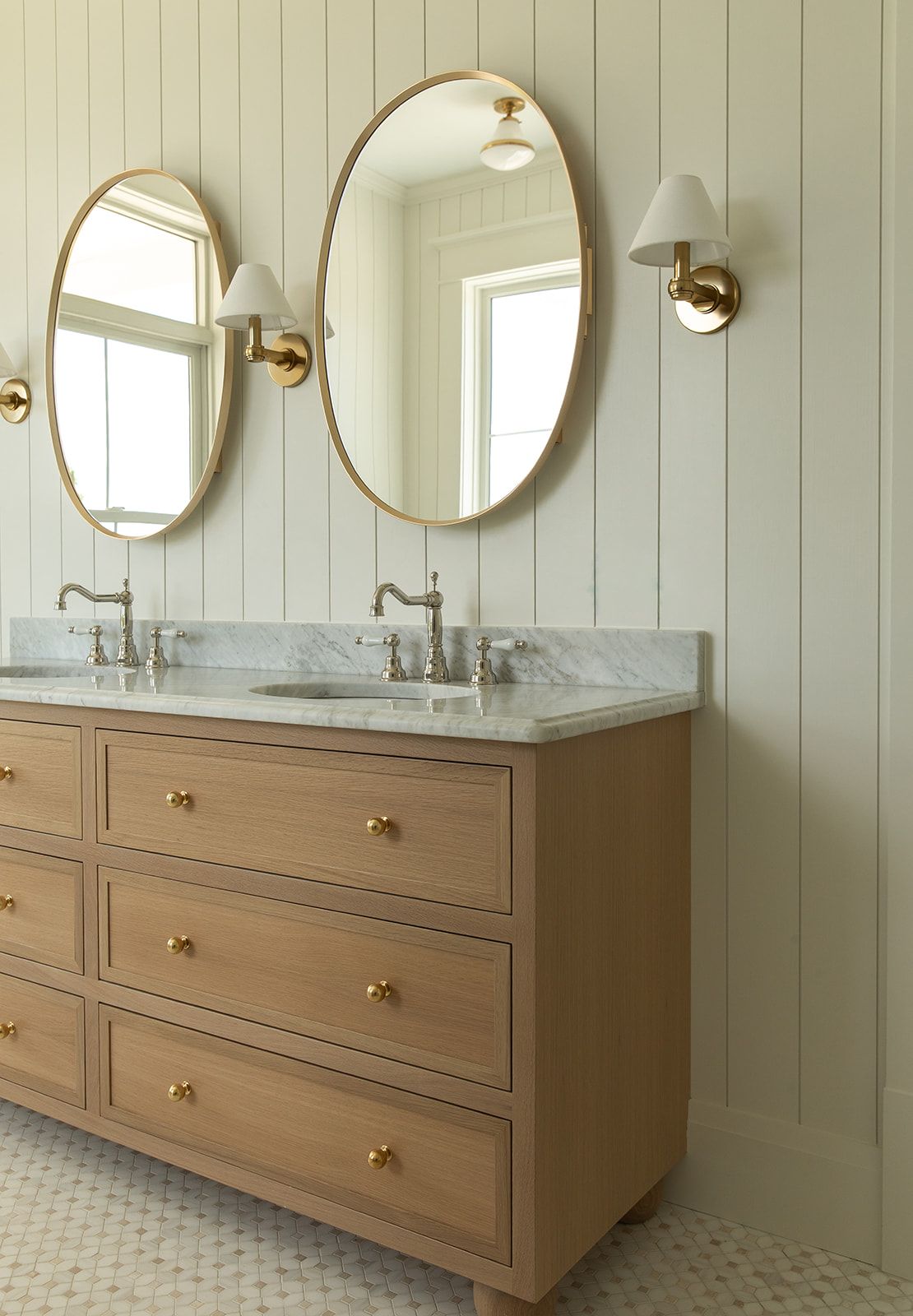 Double bathroom vanity with oak drawers, gold hardware, marble countertop, oval gold mirrors, and brass wall sconces.