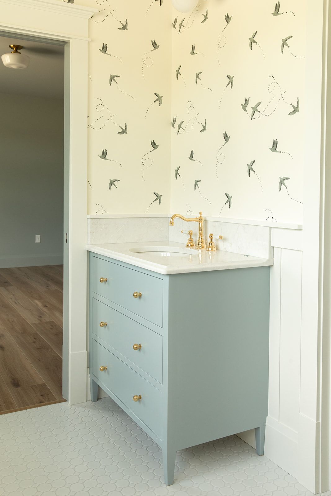 Light blue vanity with gold hardware and white marble top, paired with bird-print wallpaper above white wainscoting.