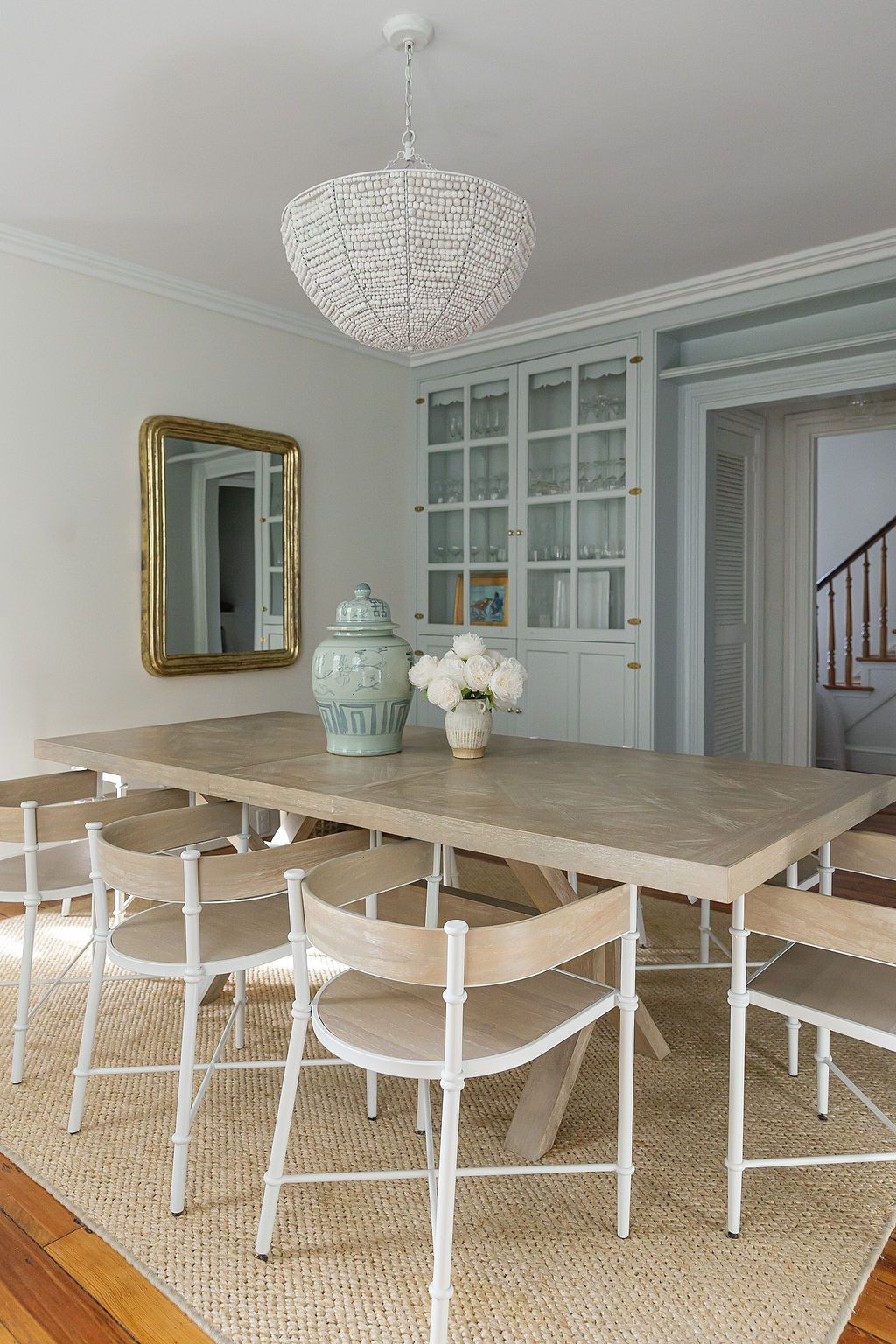 Modern coastal dining room with beaded chandelier, wood table, white chairs and blue-green built-in cabinet on sisal rug.