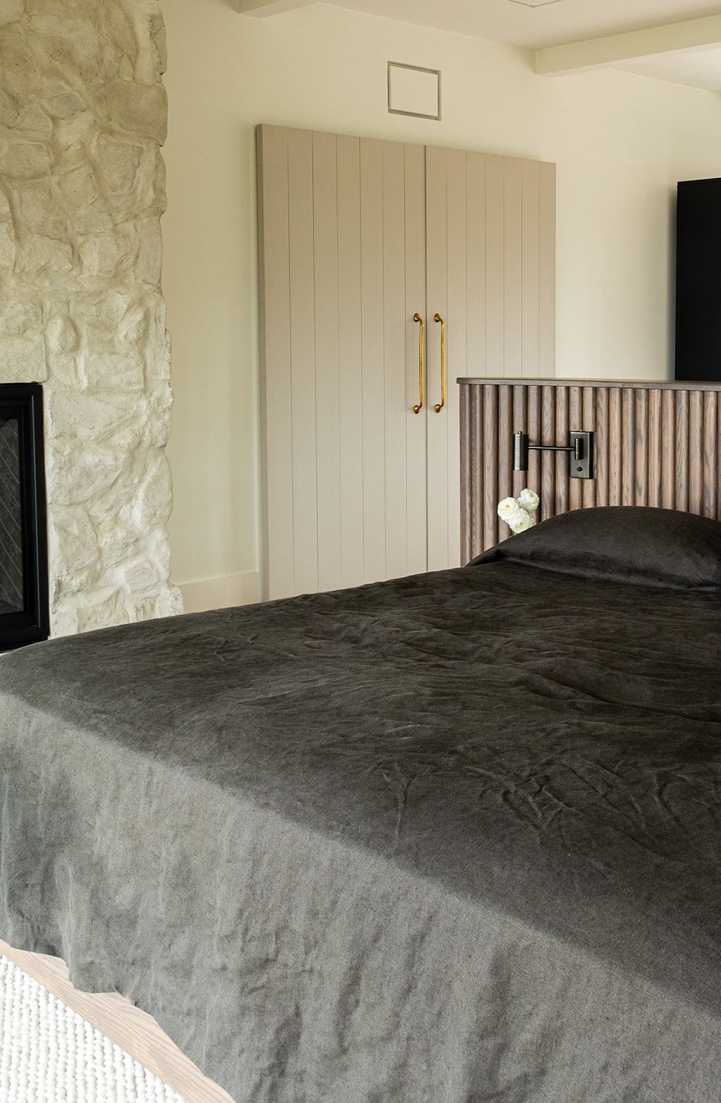 Modern bedroom with dark olive linen bedding, stone fireplace, beige wardrobe with gold handles, and slatted headboard.