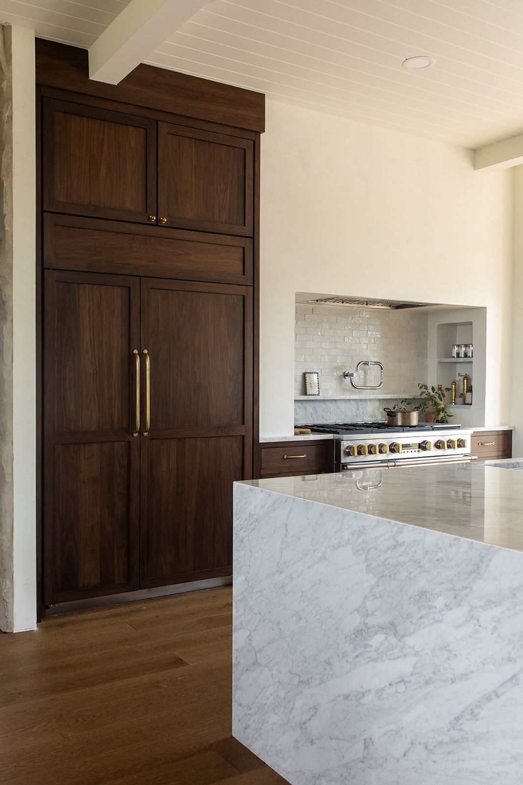 Dark walnut panel-front refrigerator with brass handles beside a marble kitchen island and stainless range.