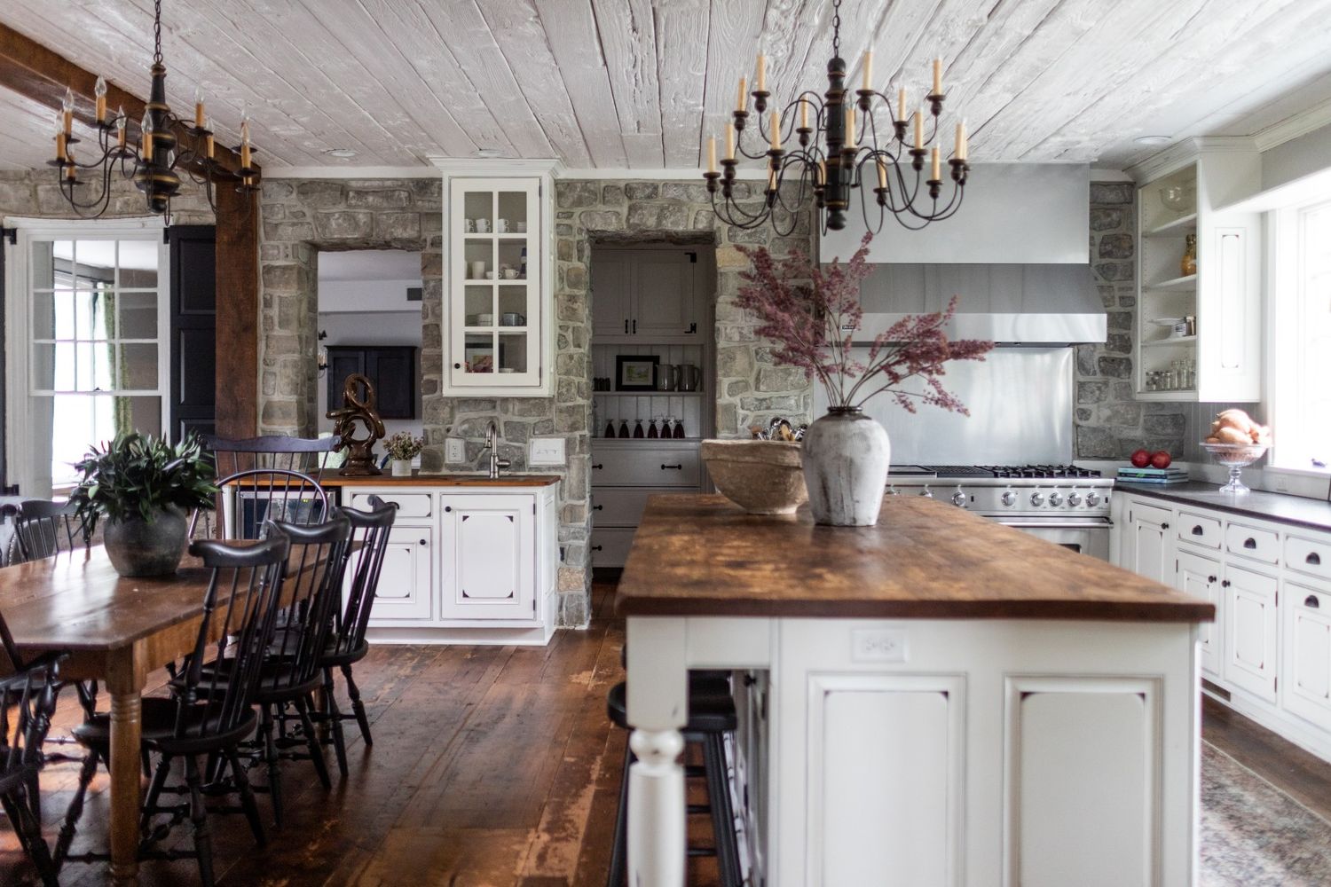 Rustic farmhouse kitchen with white cabinets, wood countertops, stone walls, and elegant chandeliers hanging from a wood ceiling.
