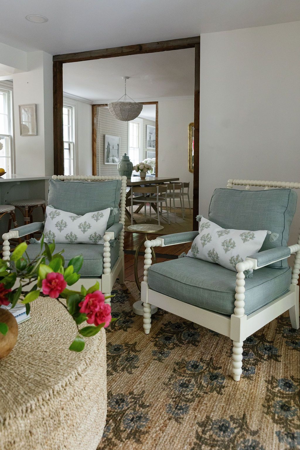 Elegant living room with seafoam blue upholstered chairs, floral pillows, and a round woven ottoman near a large mirror.