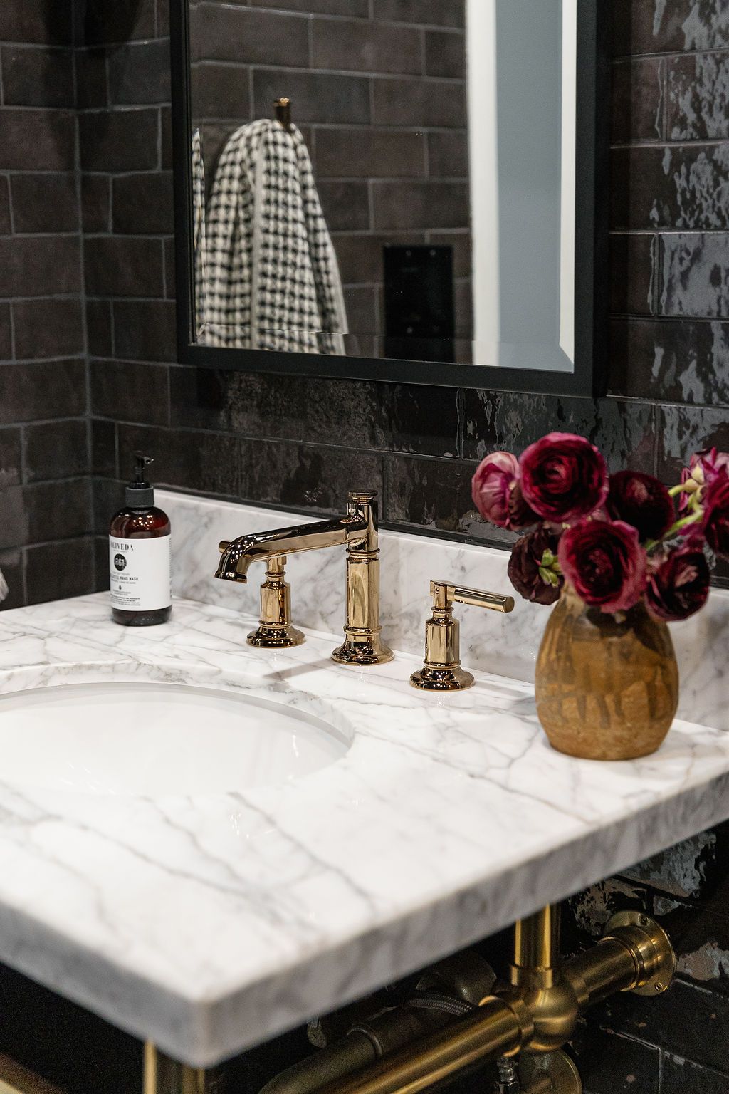 Elegant bathroom vanity with marble countertop, gold fixtures, dark tile walls, and burgundy roses in amber vase.