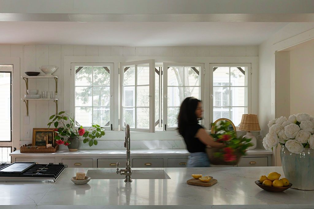 Modern white kitchen with arched windows and sunlight streaming in, featuring a long countertop with fresh fruits and plants.