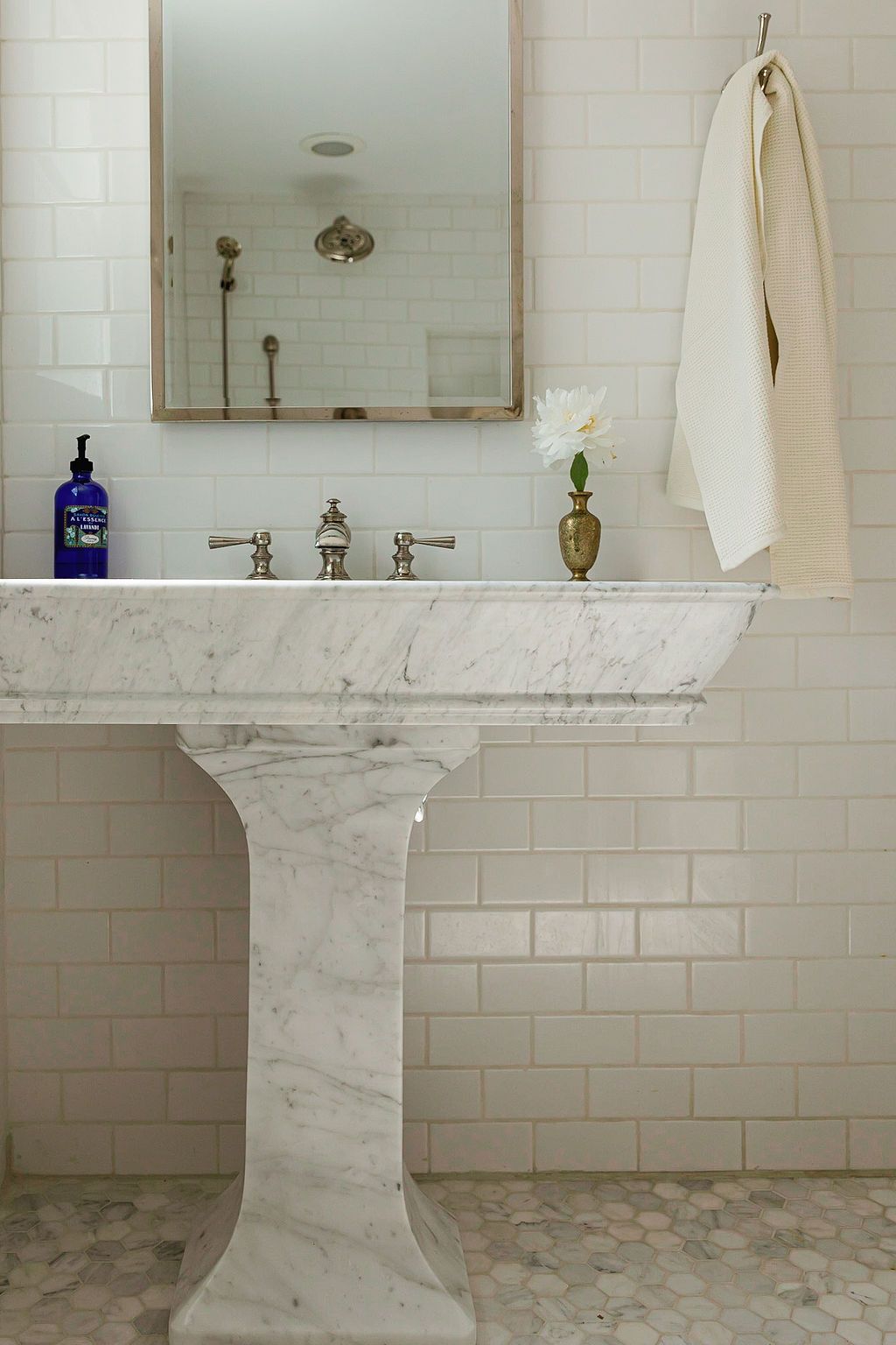 White marble pedestal sink with vintage chrome faucet sits against subway tile wall beneath framed mirror and blue glass bottle.
