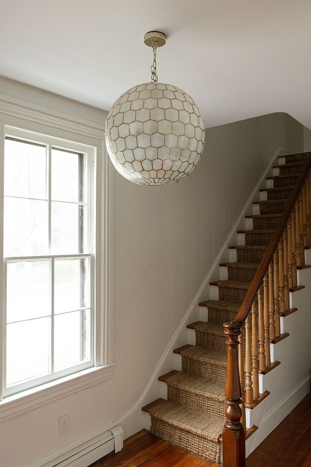 A spherical capiz shell pendant light hangs above a carpeted staircase with wooden banister in a bright interior space.