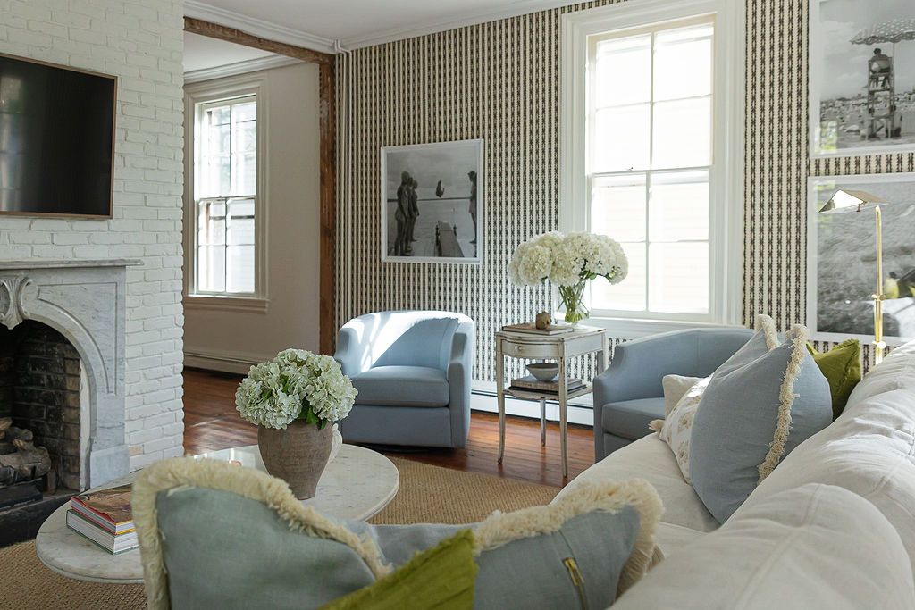 A modern living room with pale blue armchairs, patterned wallpaper, tall windows and a white stone fireplace.