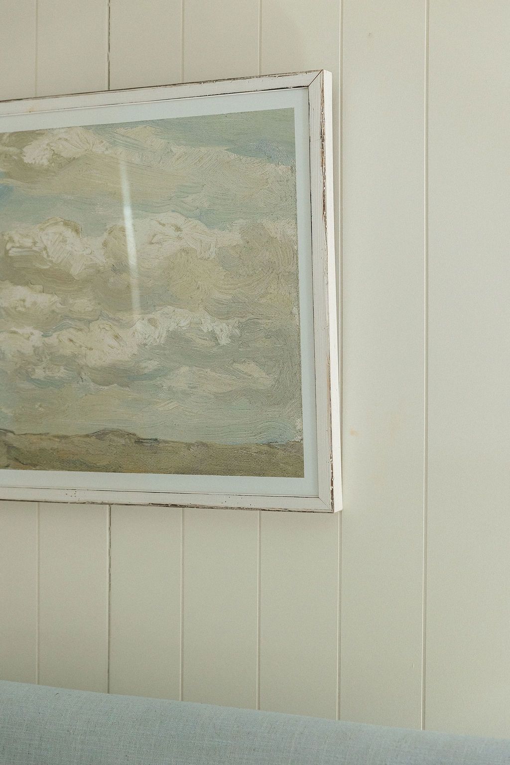 A framed landscape painting of cloudy skies hangs on a white paneled wall above a light blue surface.