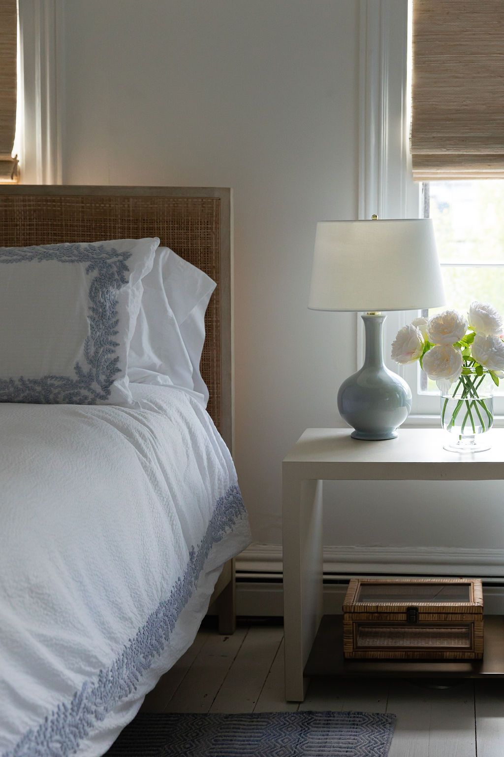 Minimalist bedroom corner featuring white bedding, woven headboard, and soft blue table lamp by window.