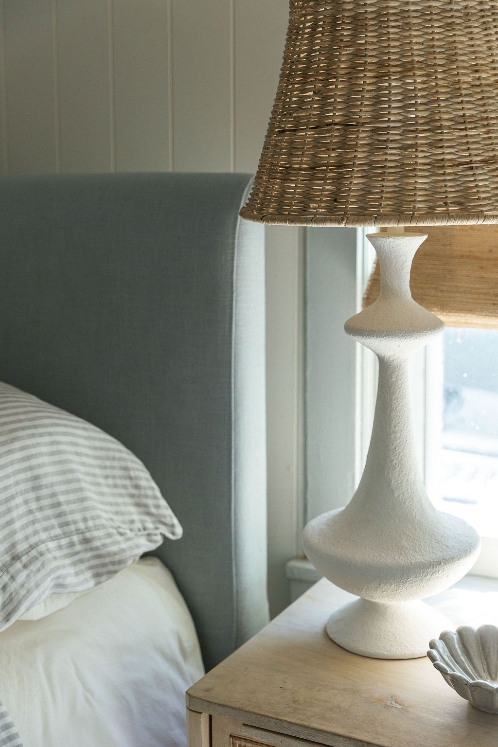 A white ceramic table lamp with woven basket shade sits on a wooden nightstand beside a striped pillow and upholstered headboard.