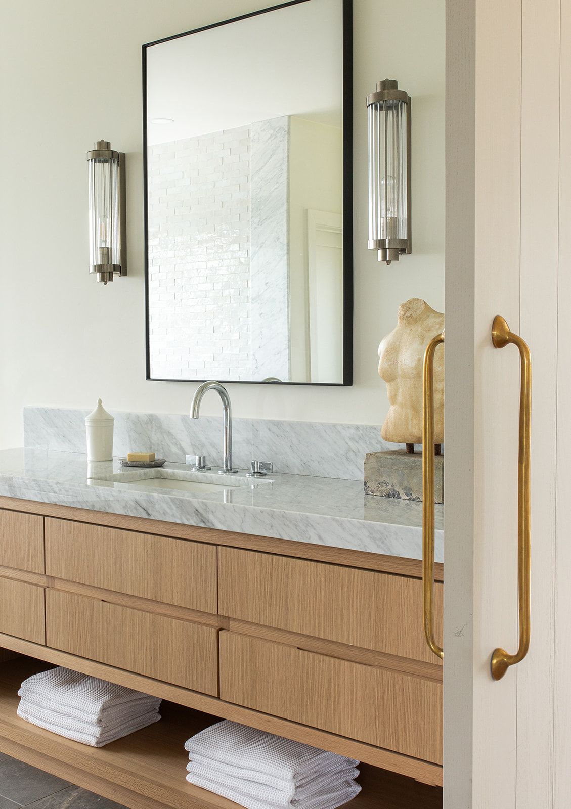 Light oak bathroom vanity with marble countertop, black framed mirror, crystal sconces, and gold door pull.