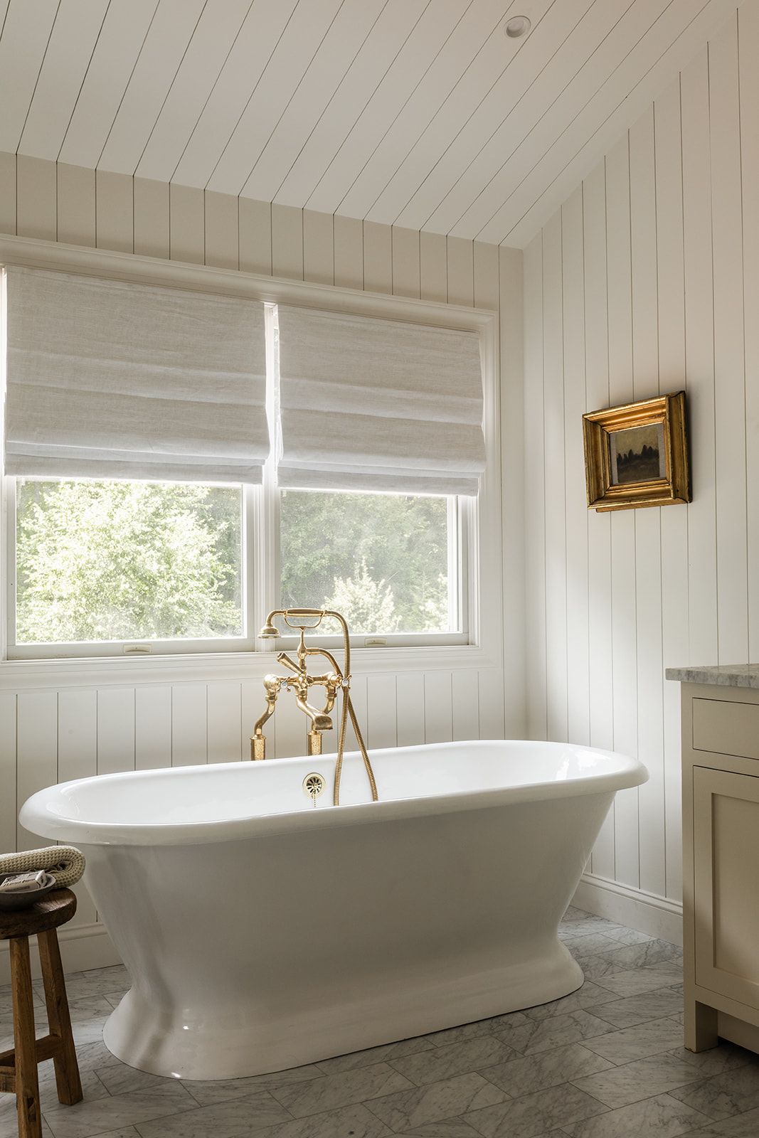 White freestanding bathtub with gold fixtures in a bright bathroom featuring shiplap walls, Roman shades, and a small landscape painting.