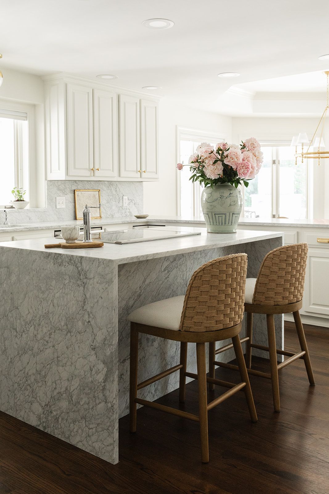 White kitchen with marble island, woven counter stools, pink peonies, and gold hardware accents on white cabinetry.