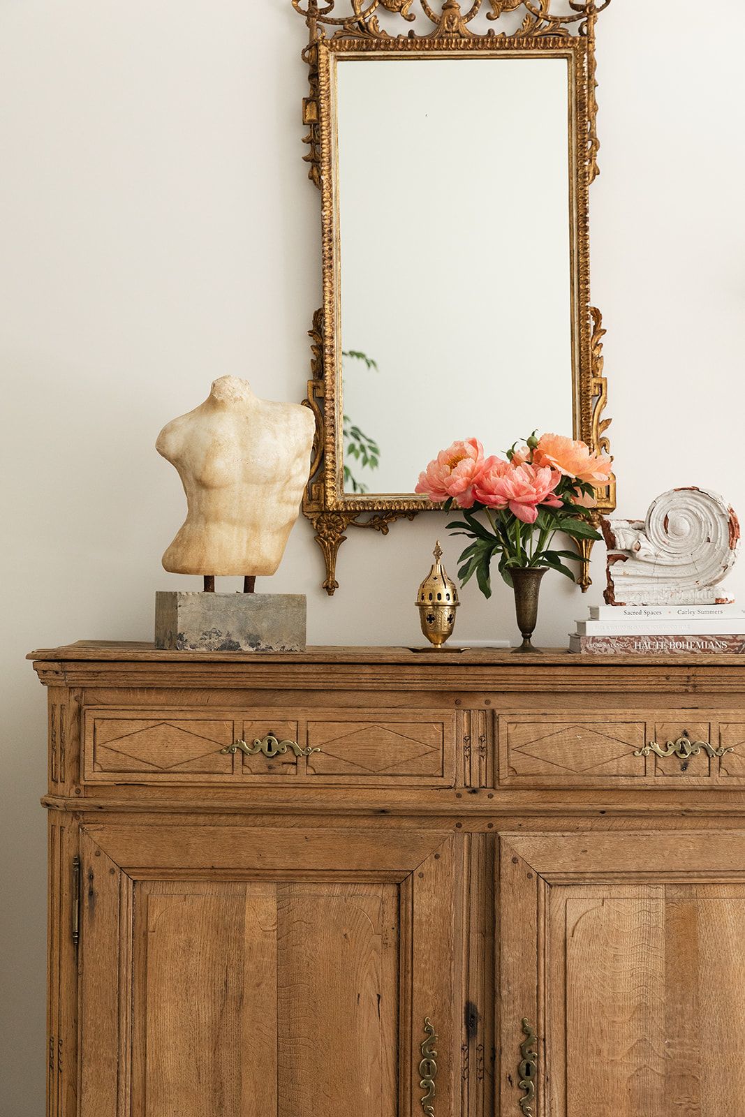 Antique wooden sideboard with gold ornate mirror, pink peonies, marble torso sculpture, and decorative accents.
