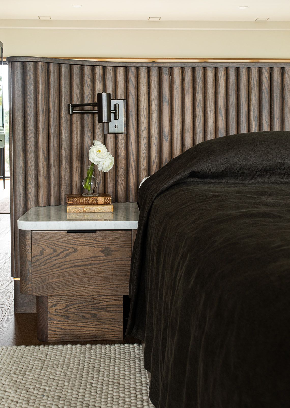 Modern bedroom with dark wood fluted wall panel, black bedding, marble nightstand with books and white flower, and swing arm sconce.