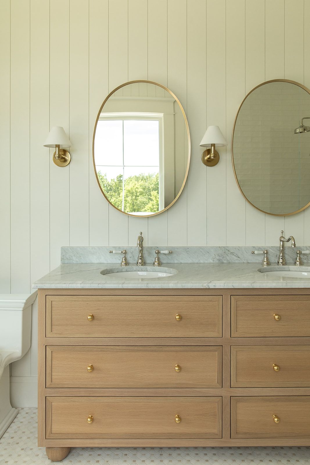 Elegant double sink vanity featuring light oak cabinetry, marble top, gold oval mirrors, and white wall sconces.