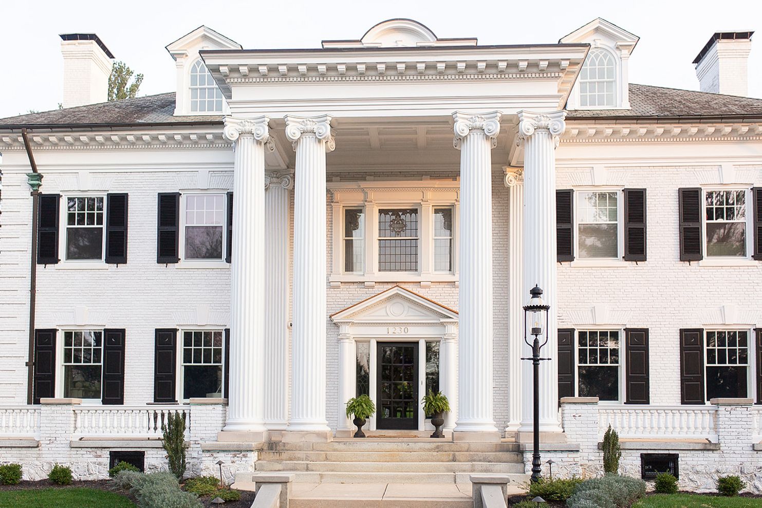 Grand white Colonial mansion with black shutters, tall columns, and elegant entrance steps surrounded by manicured landscaping.