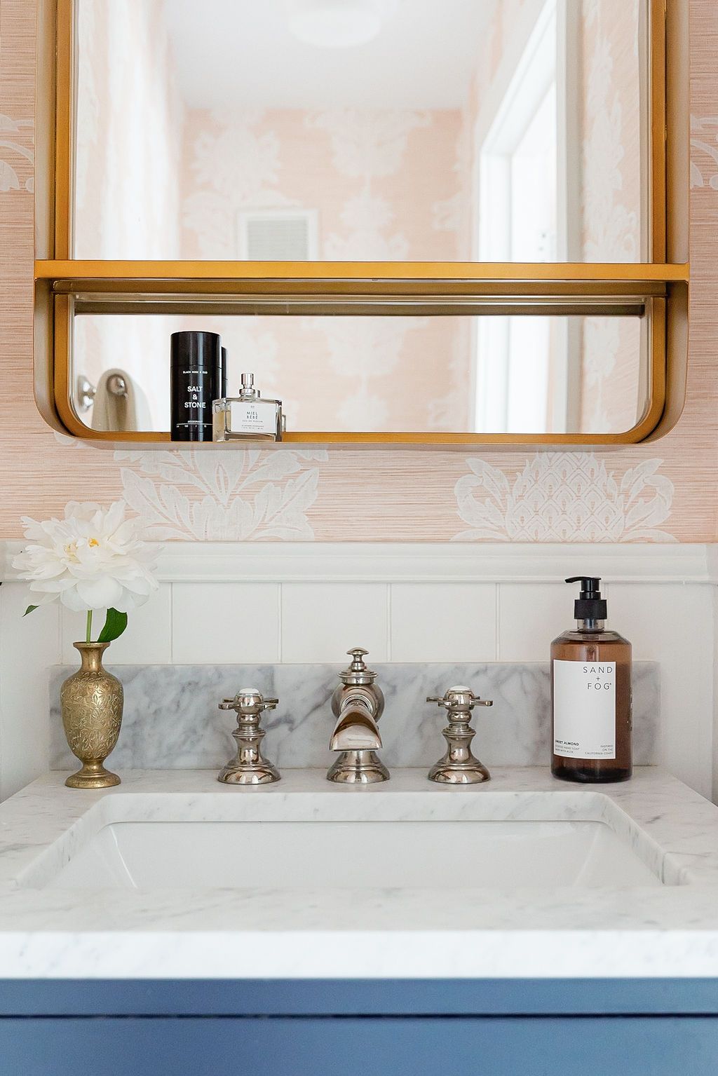 Elegant bathroom vanity with marble countertop, brass fixtures, and wood-framed mirror shelf featuring toiletries.