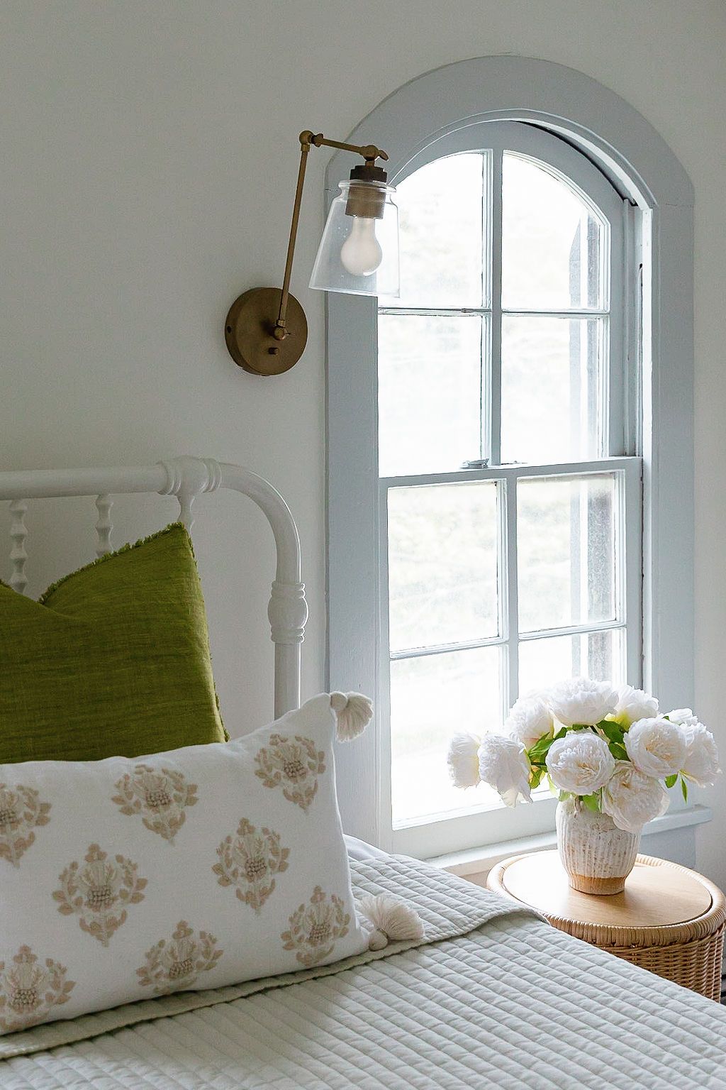 Cozy bedroom corner with arched window, white bedding, green accent pillow, and fresh peonies on side table with wall sconce.