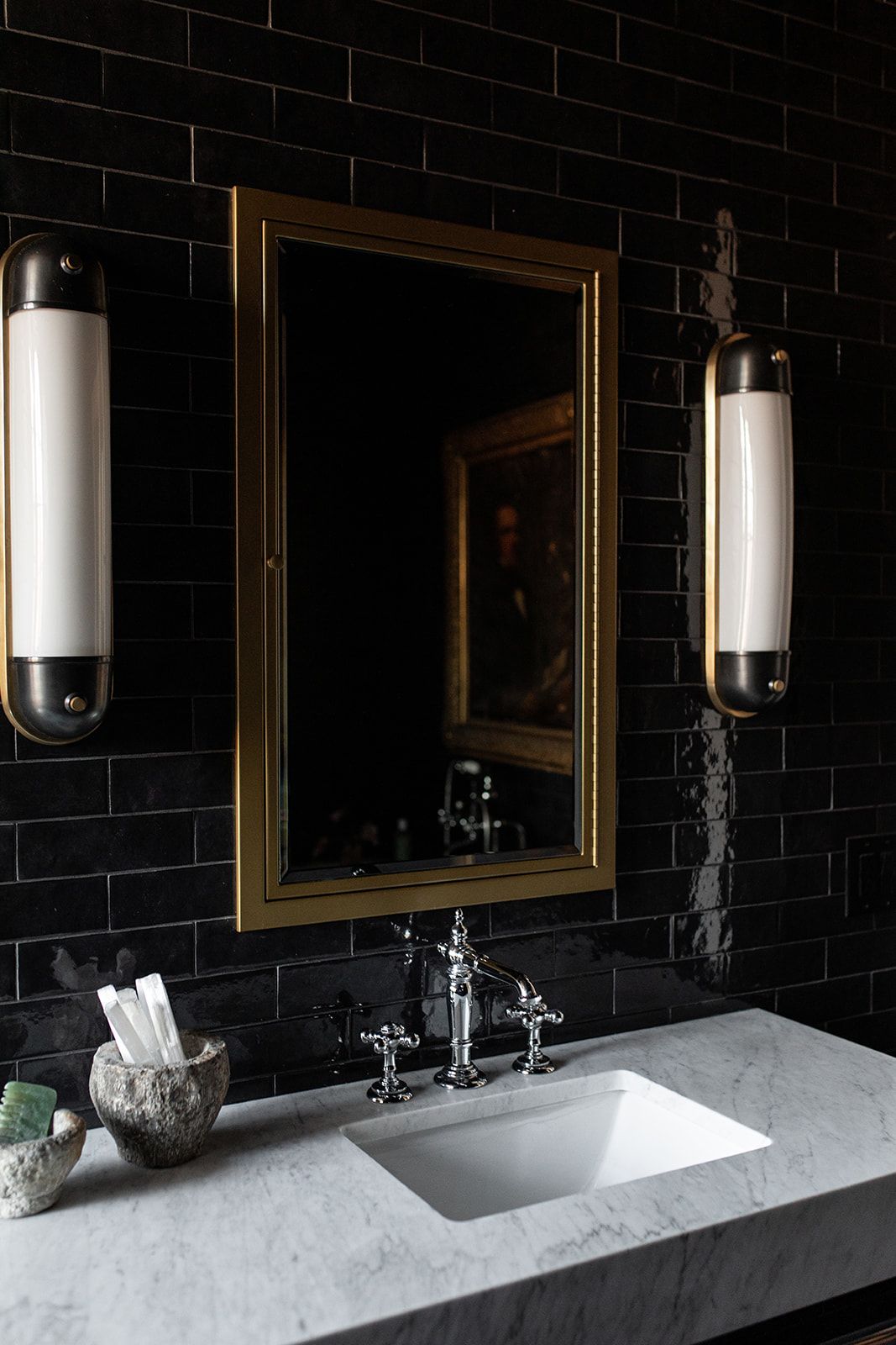 Dramatic bathroom featuring black tile walls, gold-framed mirror, and marble vanity with sconce lighting.