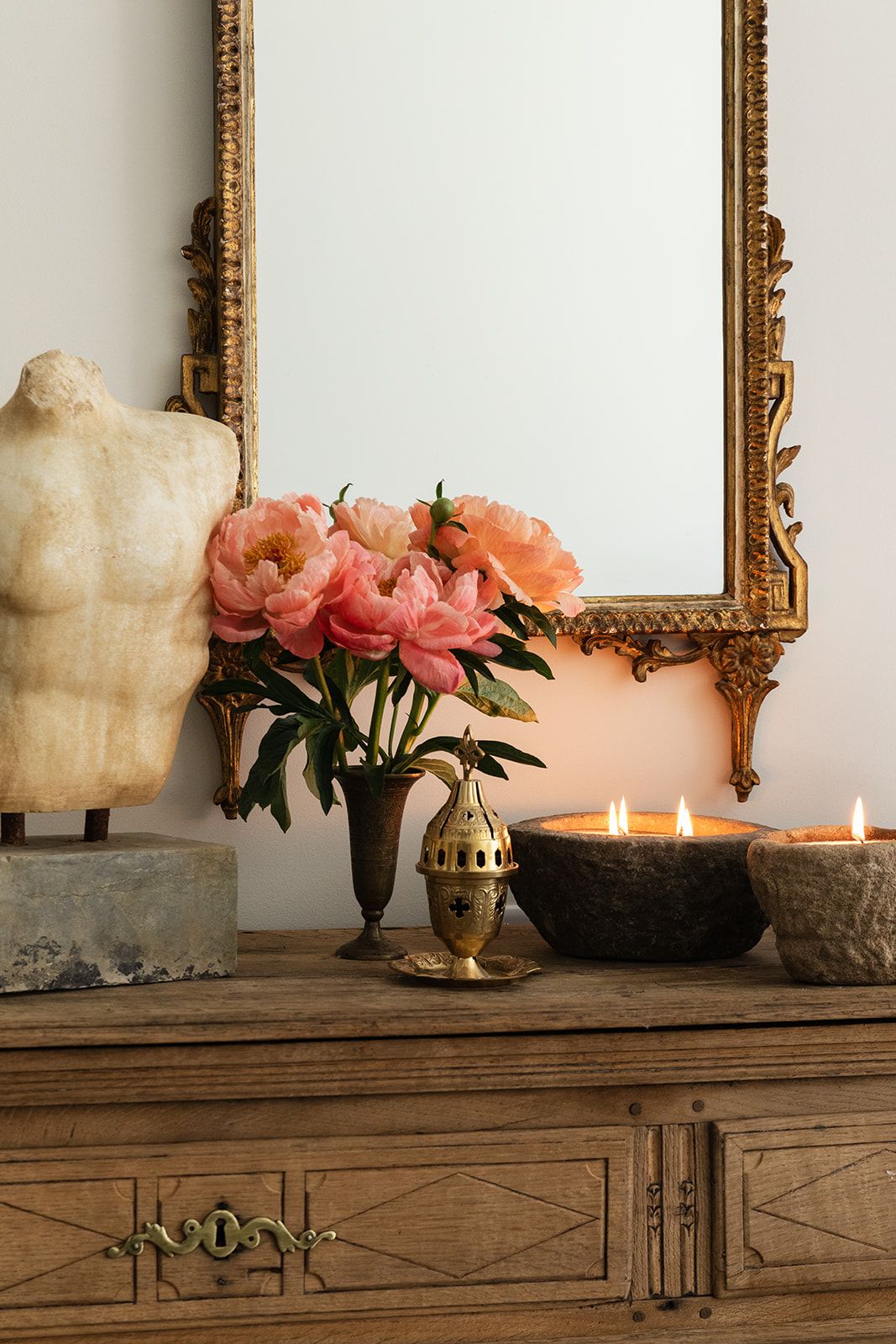 Ornate gold mirror above wooden dresser with pink peonies, lit candles, brass vase, and classical torso sculpture.