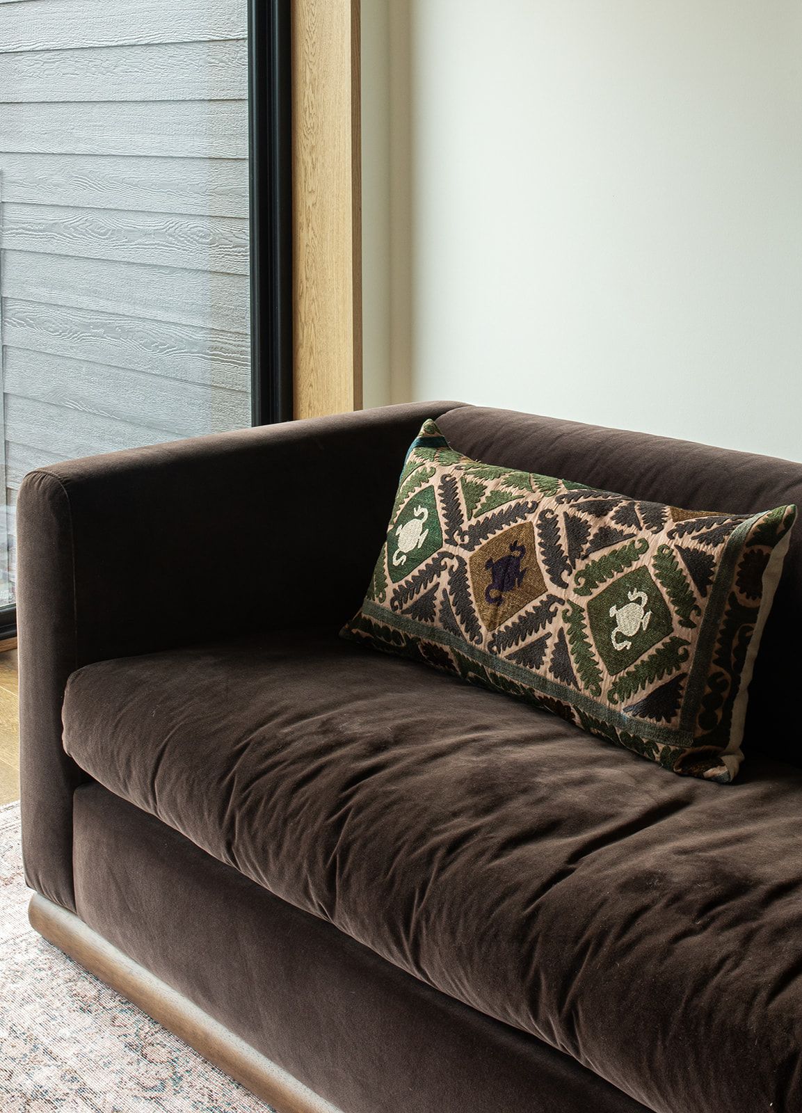 Dark brown velvet loveseat with decorative geometric embroidered lumbar pillow in green and neutral tones.