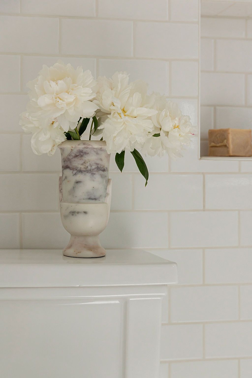 White peonies in a marbled ceramic vase against white subway tile wall on a white surface.