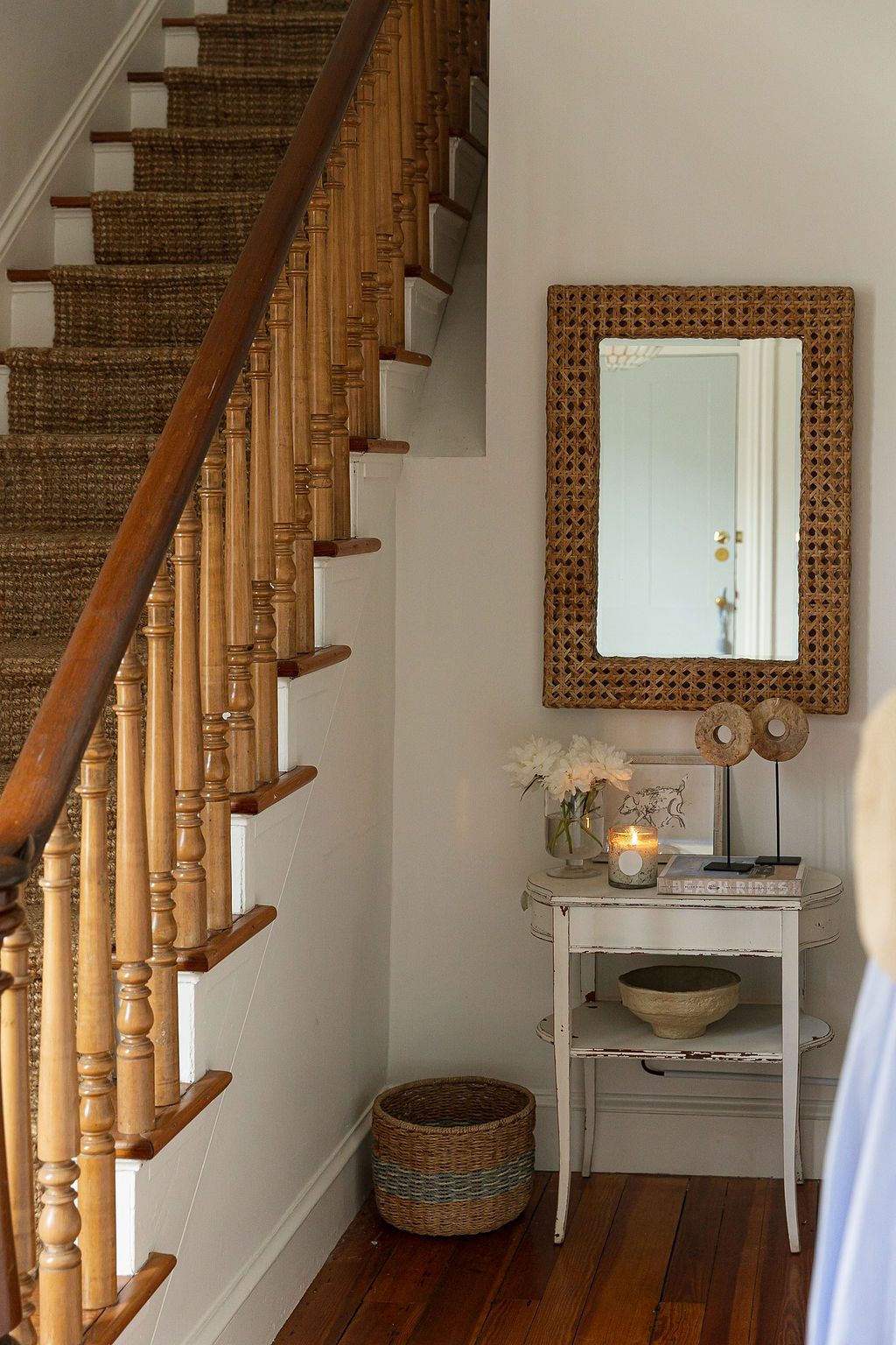 Charming home entryway with wooden staircase, woven mirror, small white console table and decorative basket beside stairs.